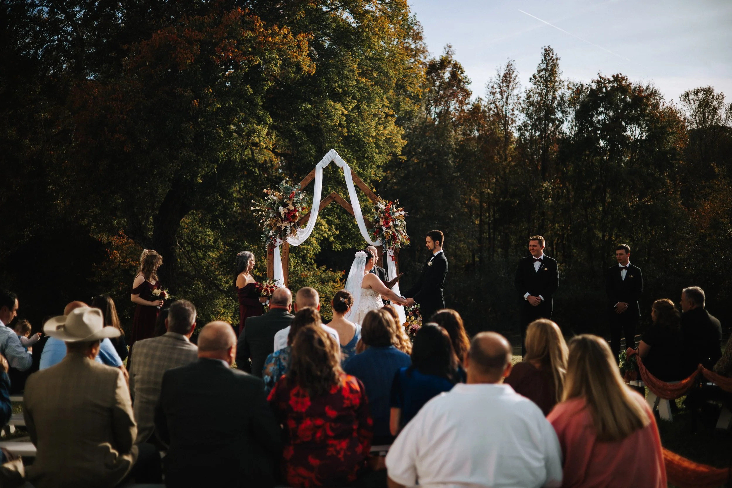 A wedding ceremony taking place outdoors in a park, with a wooden arch decorated with flowers and white fabric, where the bride and groom are exchanging vows. Guests are seated around watching the ceremony, with trees in the background and clear skie