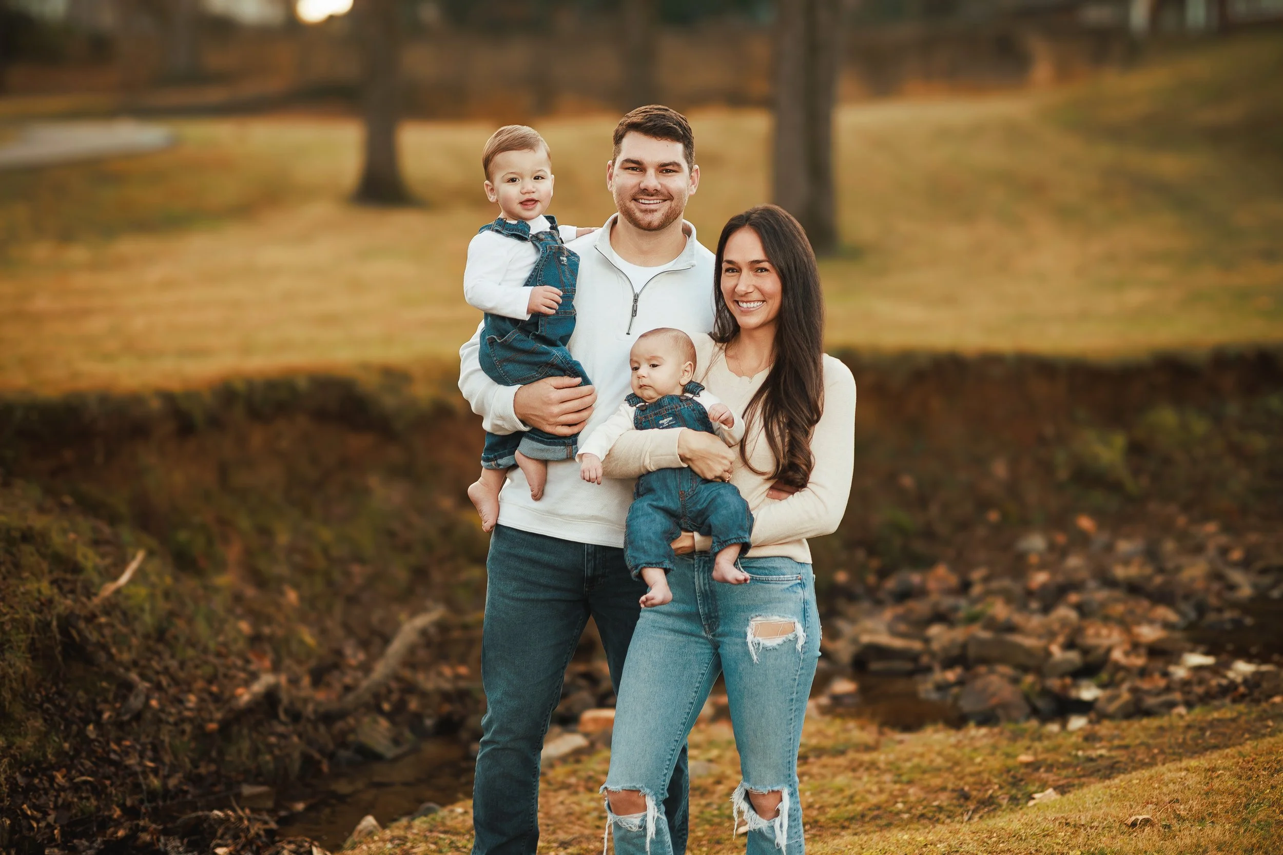 Happy family of four standing outdoors in a park during autumn, with trees and fallen leaves in the background.