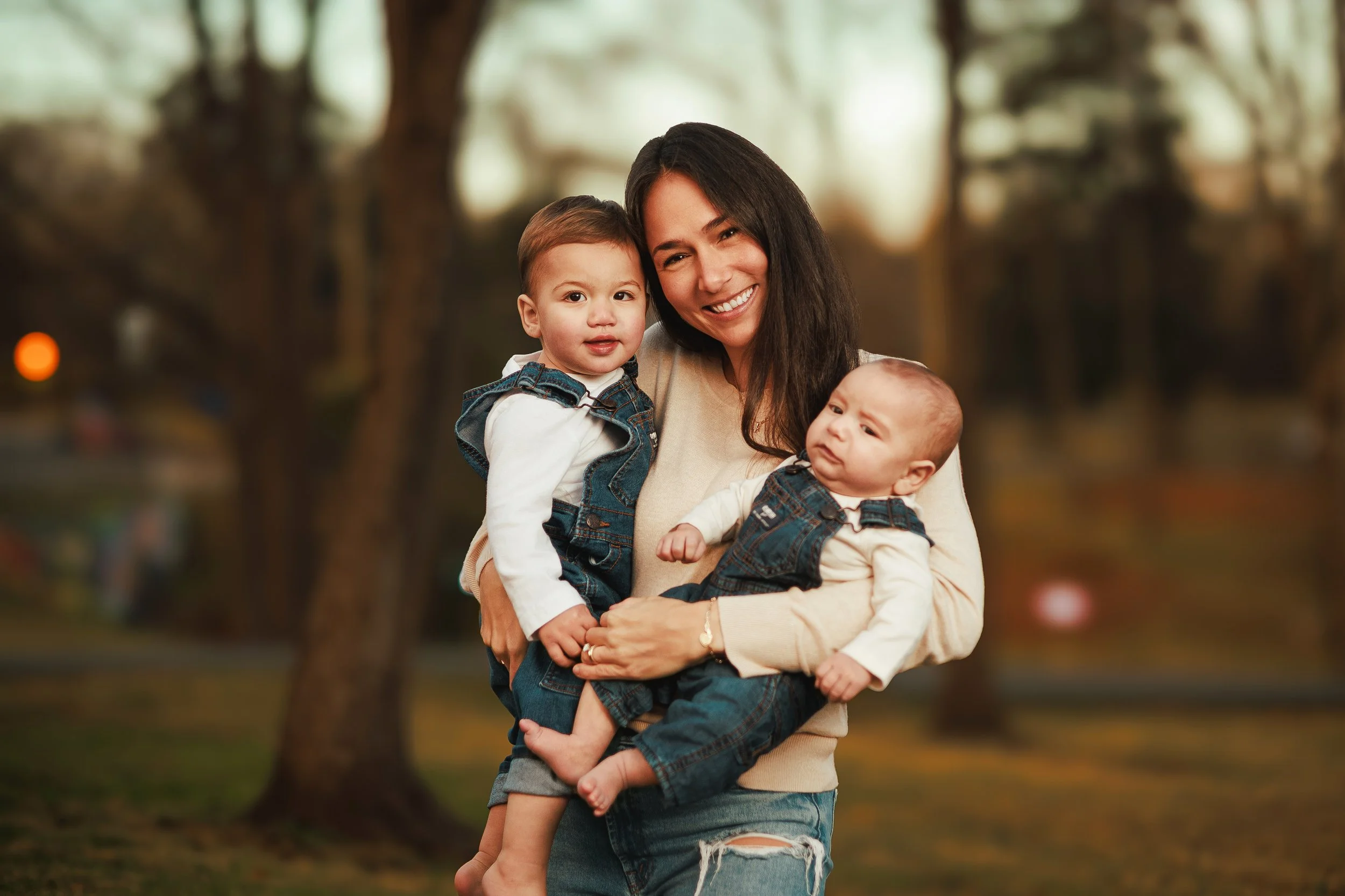 A smiling woman holding two young children outdoors in a park with trees in the background.