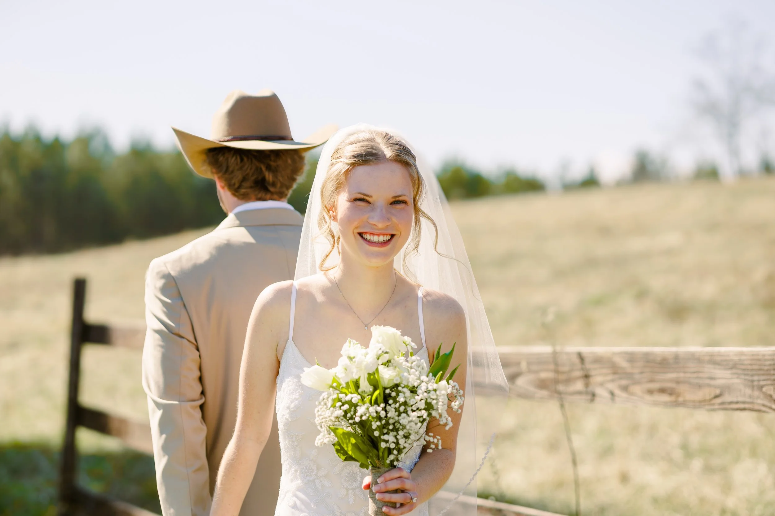 A smiling bride holding a bouquet of white flowers, standing outdoors with a groom in the background, wearing a cowboy hat, in a rural setting with a fence and trees.