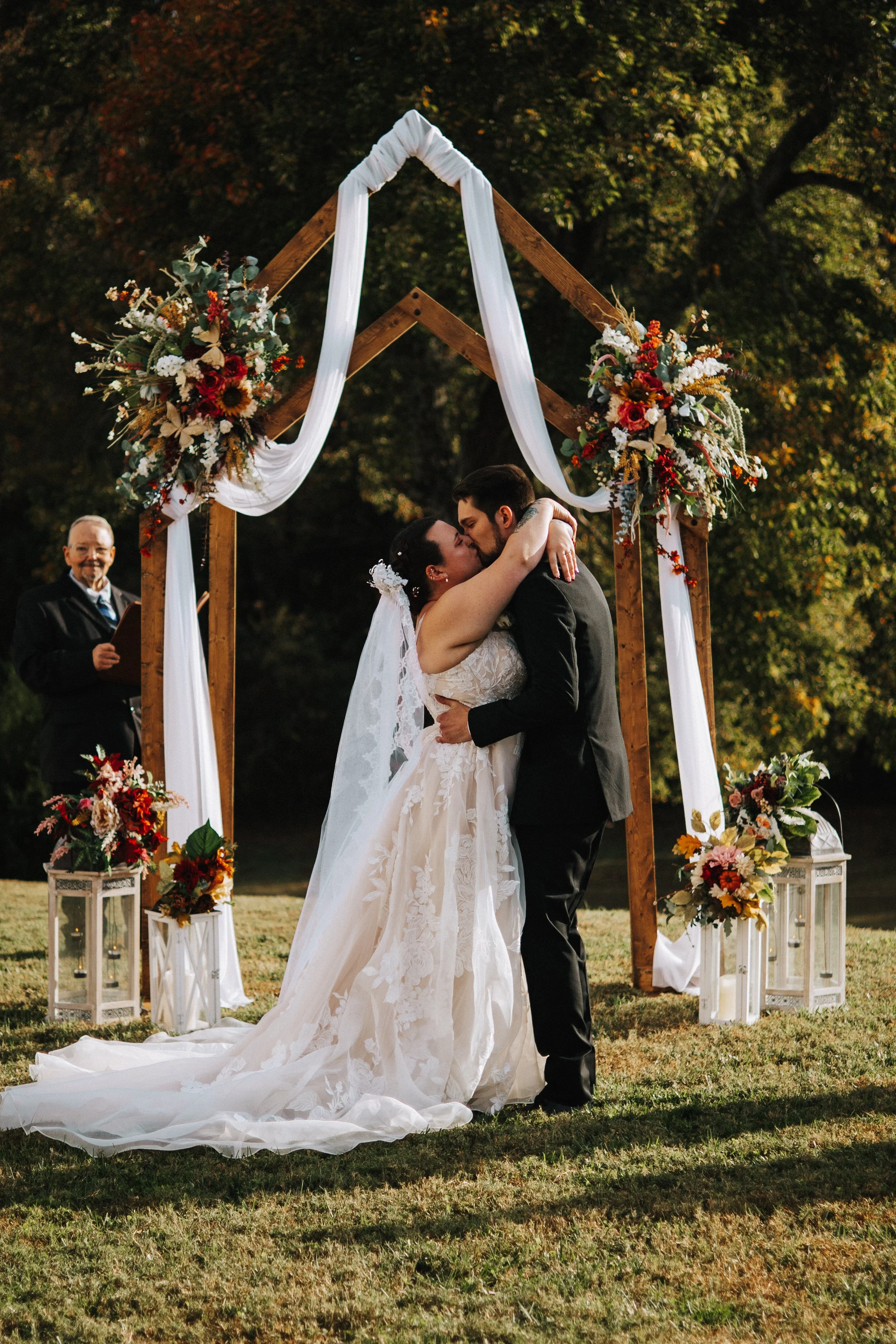 A bride and groom kissing during an outdoor wedding ceremony beneath a wooden arch decorated with flowers and white fabric, with an officiant standing nearby.