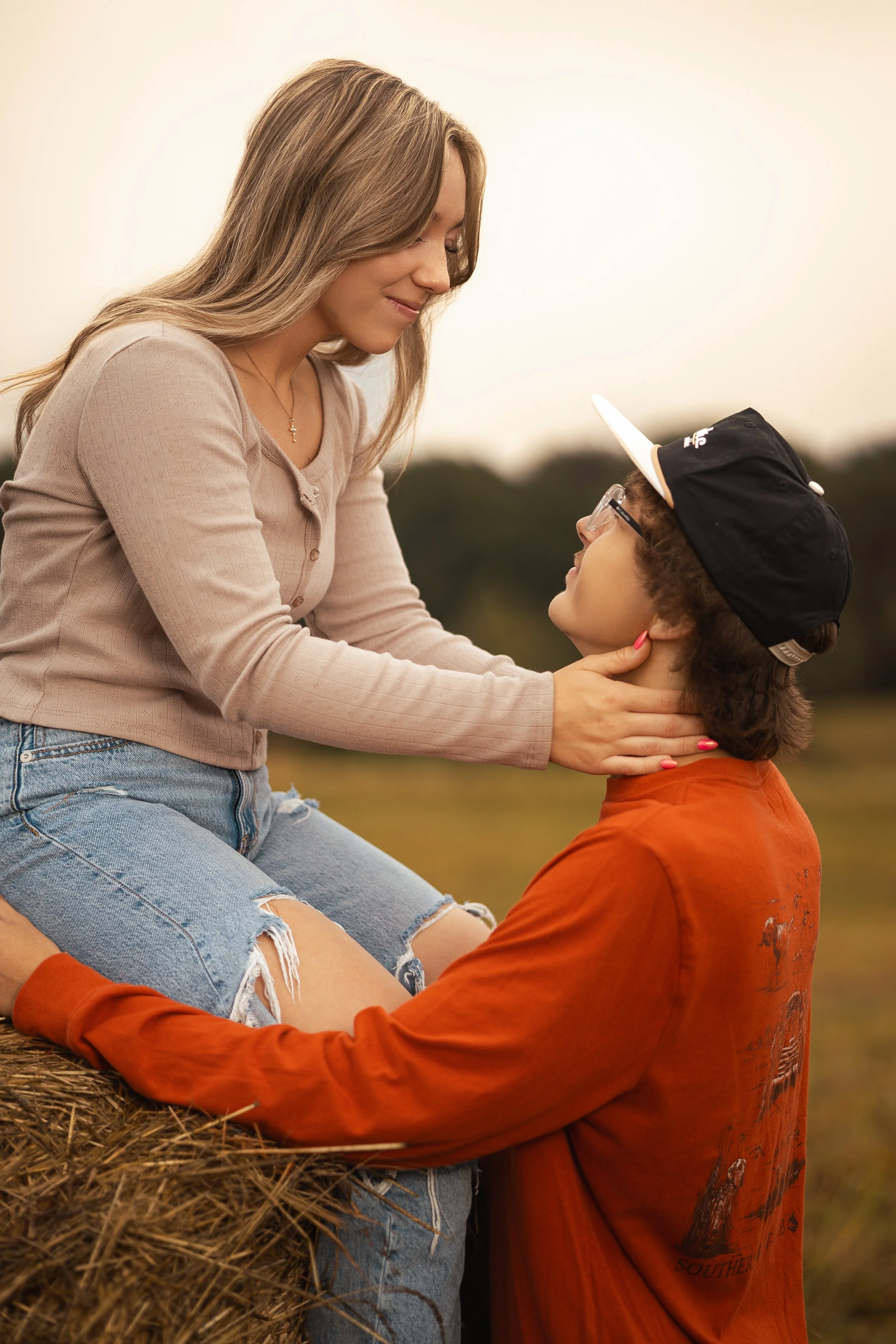 A woman with long, light brown hair and a man with curly hair, glasses, and a baseball cap sitting on a haystack outdoors, touching faces tenderly during sunset.