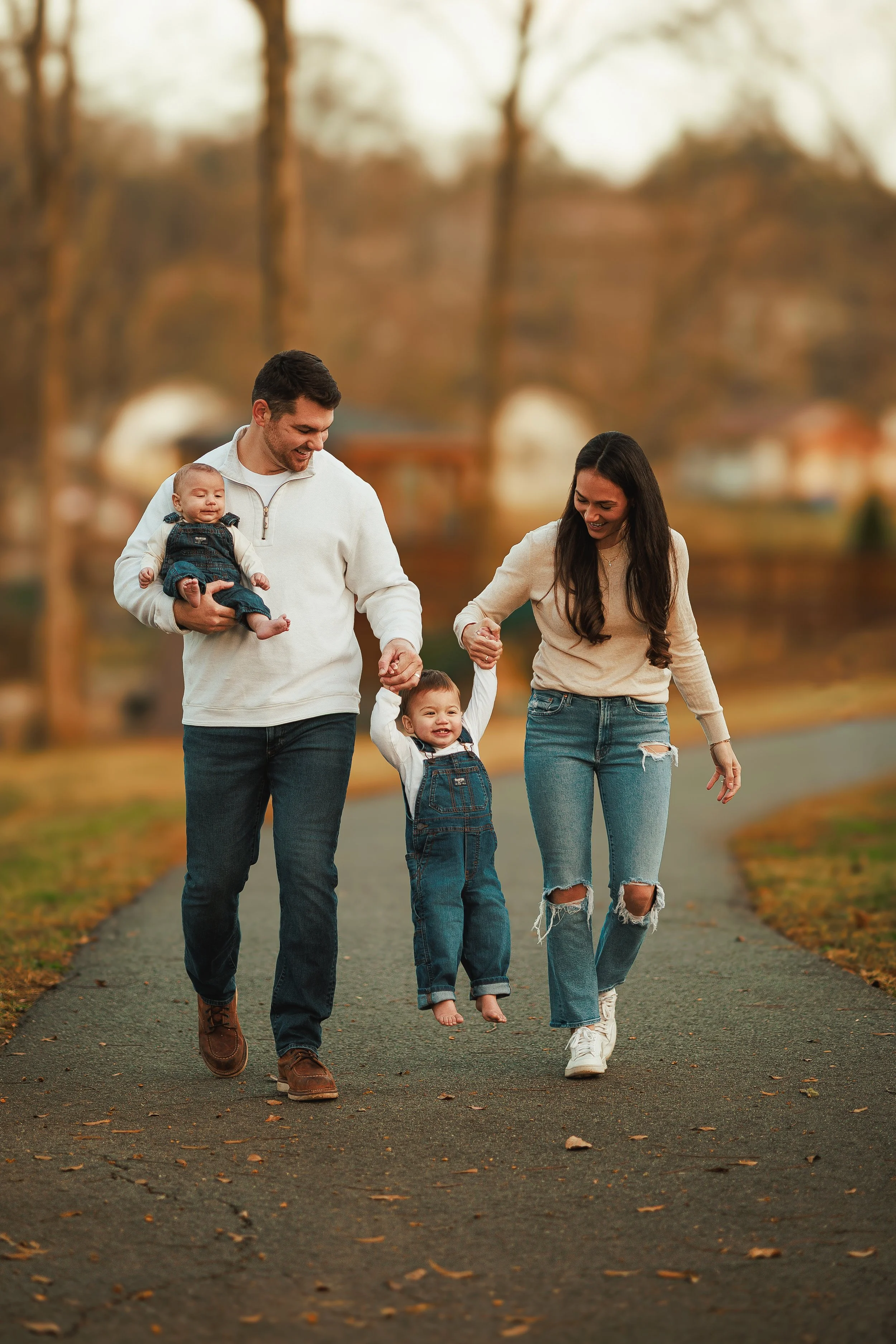 A happy family of four, including a man, woman, and two children, walking in a park during fall. The man is carrying a baby, and the woman and older child are holding hands. They are all smiling and enjoying the day.