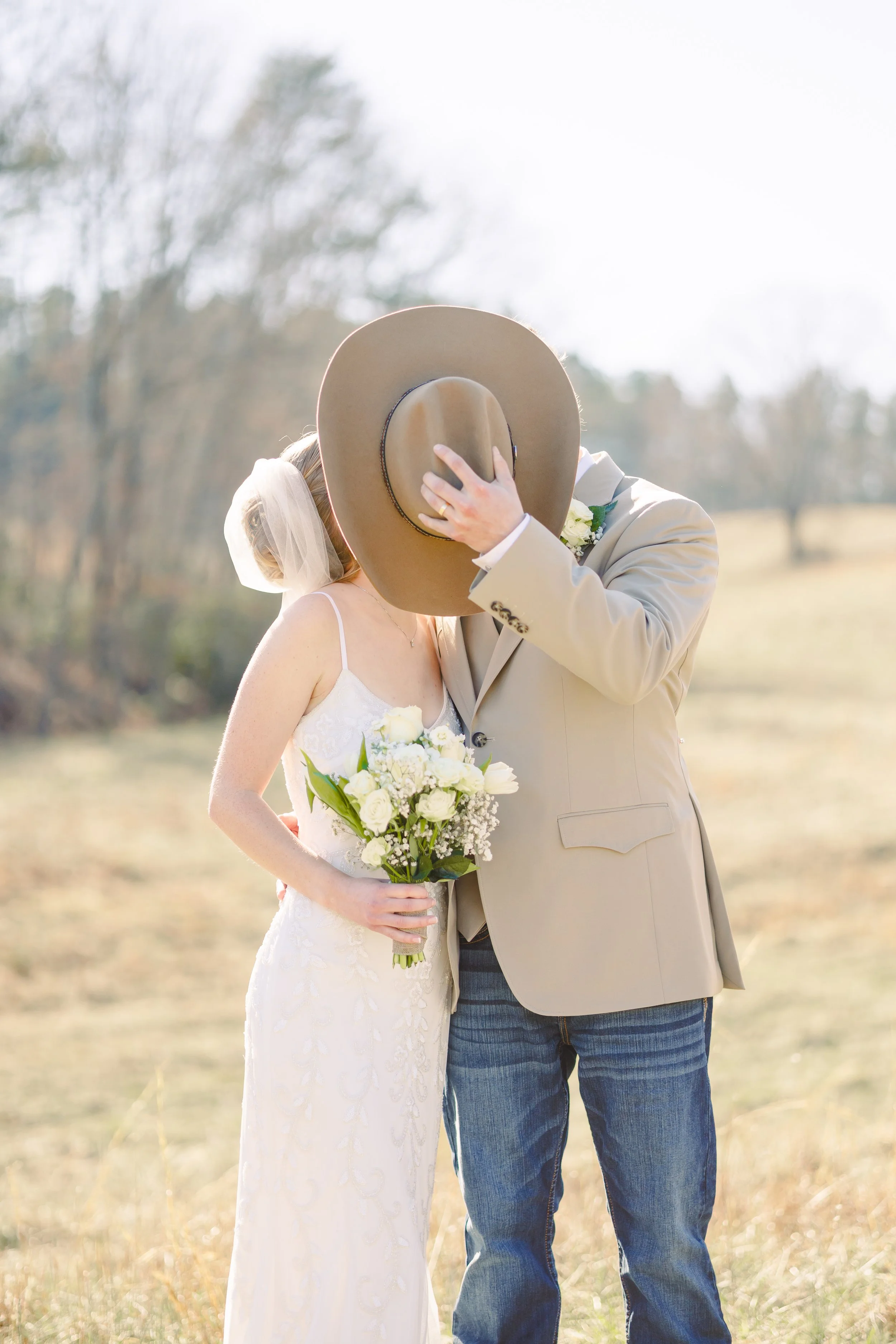 A bride and groom sharing a kiss outdoors, with the groom covering his face with a wide-brimmed hat, in a natural setting with trees in the background.