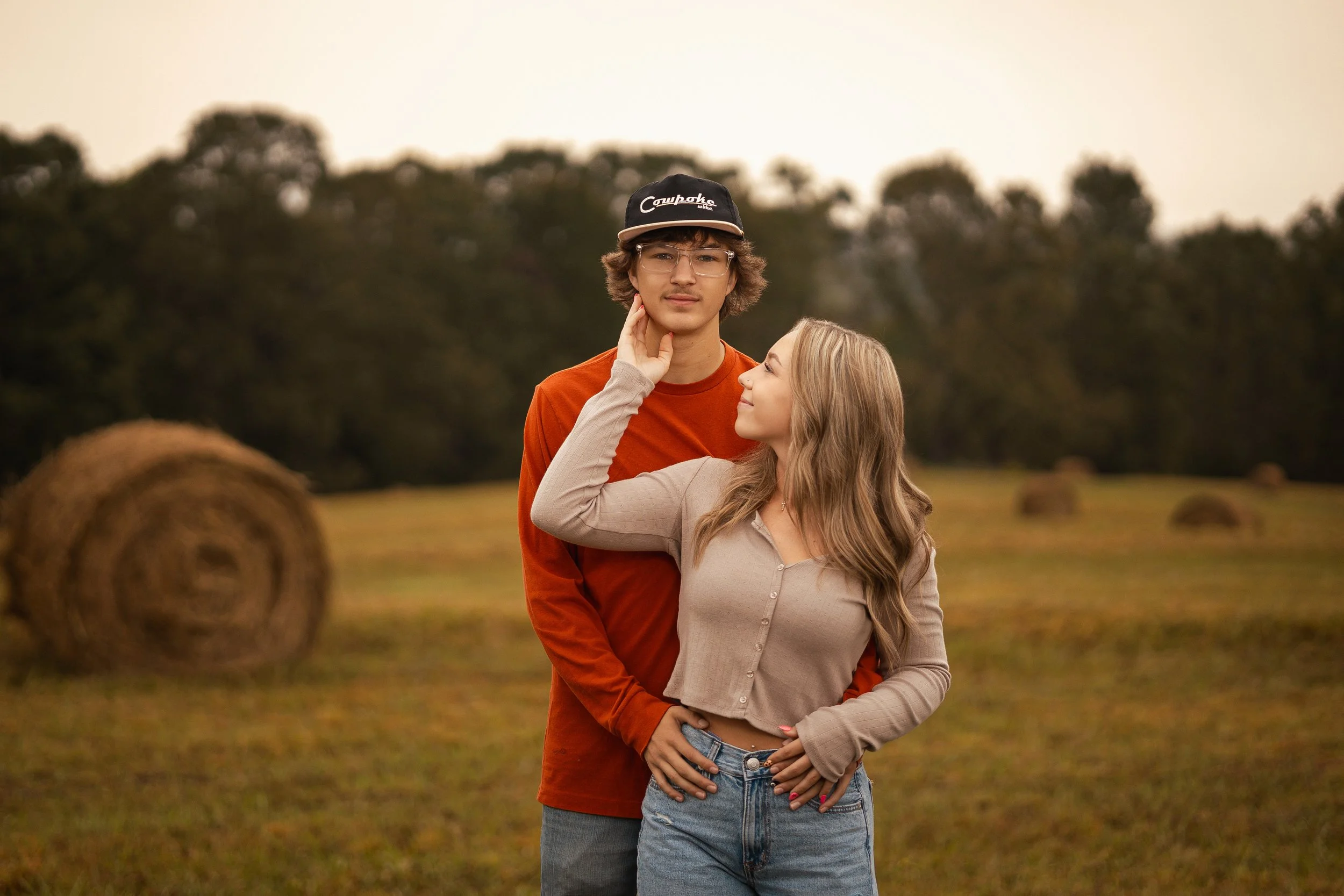 A young man and woman standing close together outdoors in a field with hay bales, with a background of trees and a cloudy sky. The woman is touching the young man's face while smiling at him.