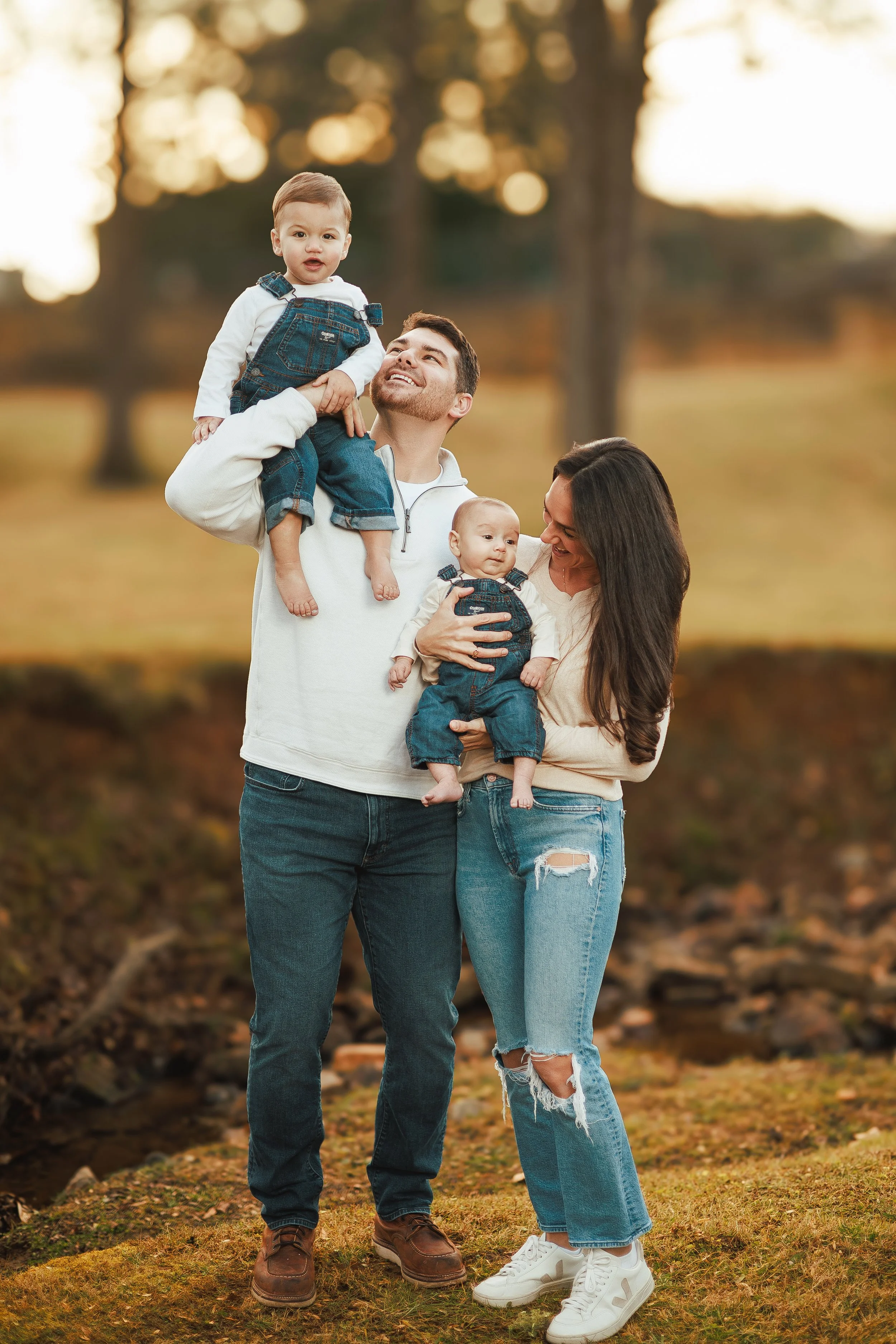 A happy family of four outdoors in a park during autumn, with trees and fallen leaves in the background. The father is holding a young boy on his shoulder, the mother is holding a baby girl, and everyone is smiling.