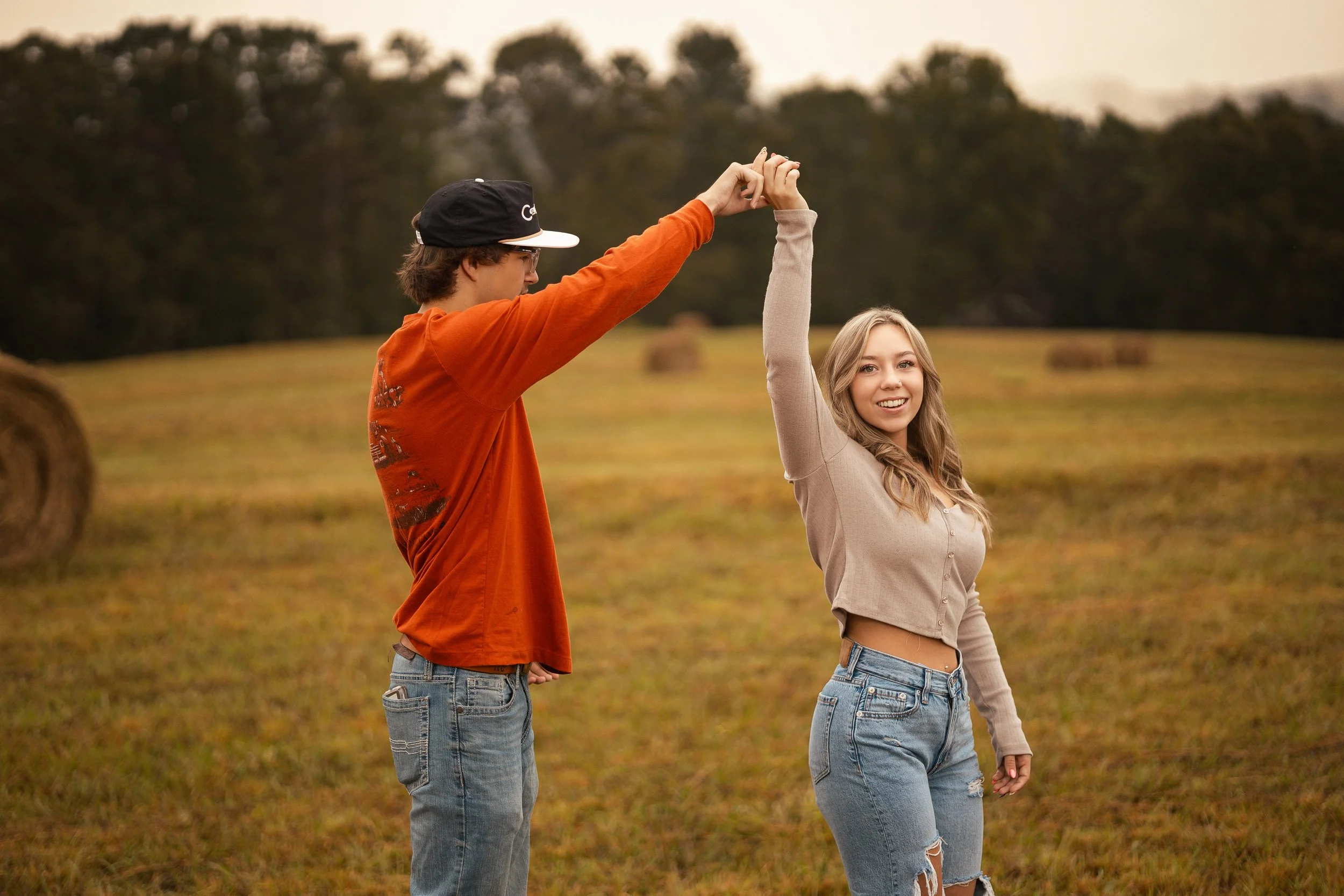 A young man and woman holding hands and waving in a field with hay bales, trees, and a cloudy sky background.