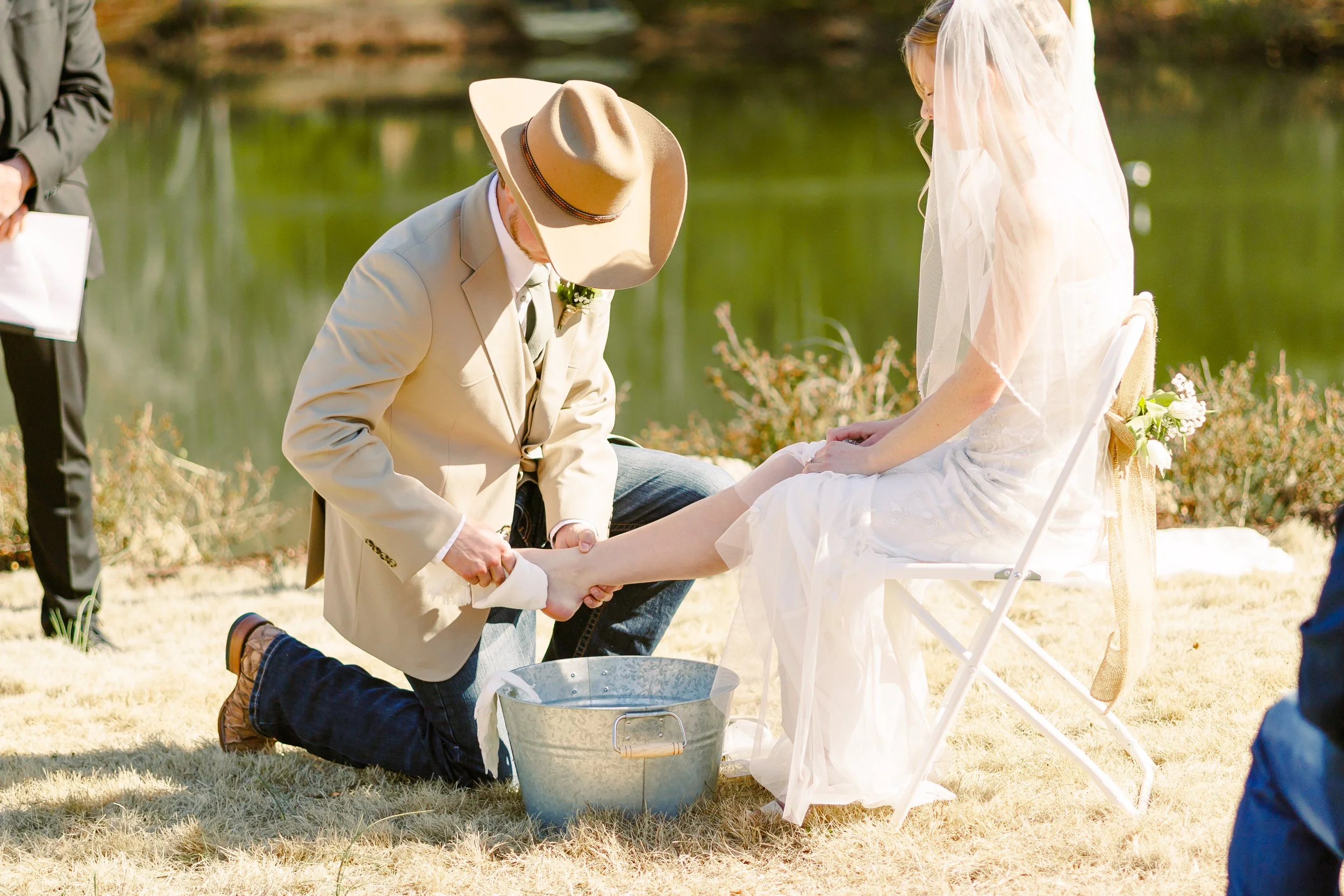 A groom kneeling and holding the bride's feet during an outdoor wedding ceremony by a lake, with the groom wearing a beige suit and large hat, and the bride sitting on a chair in a white wedding dress with a veil, holding her hands together.