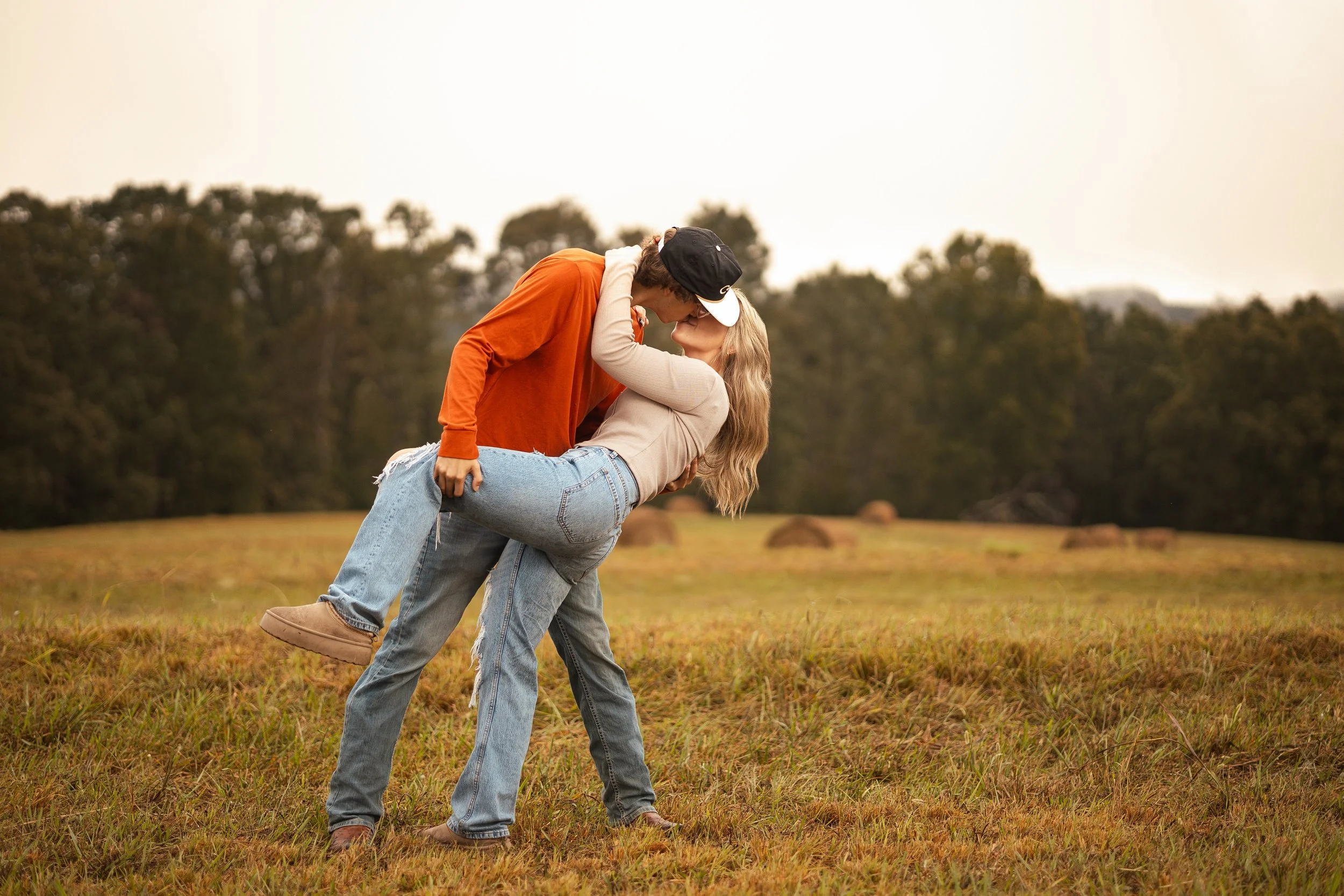 A couple sharing a kiss outdoors, with the man holding the woman in a dip against a scenic background of trees and open field.