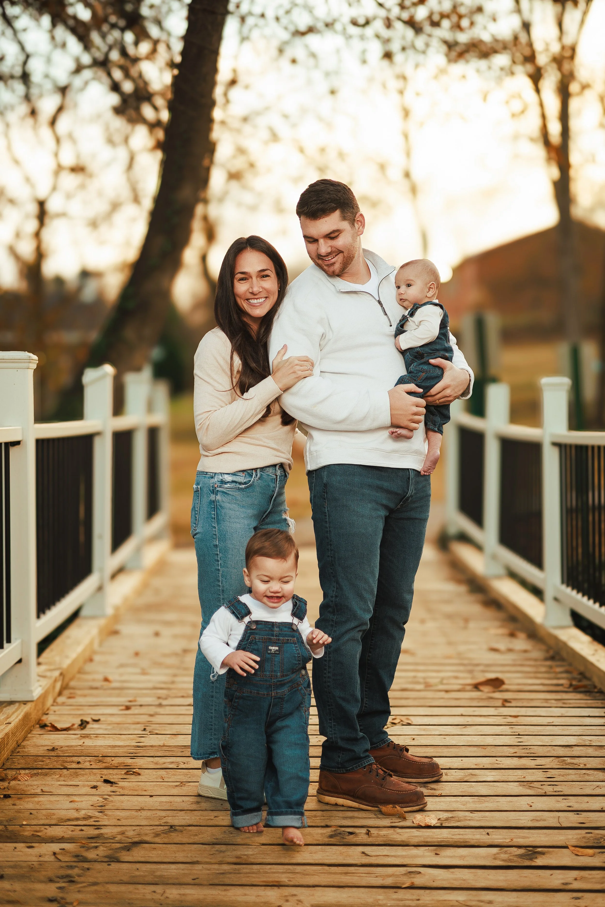 A happy family of four walking on a wooden bridge outdoors during autumn. The mother and father are smiling, with the mother hugging the father from behind as they carry their two young children, one older child and one toddler, all dressed in casual