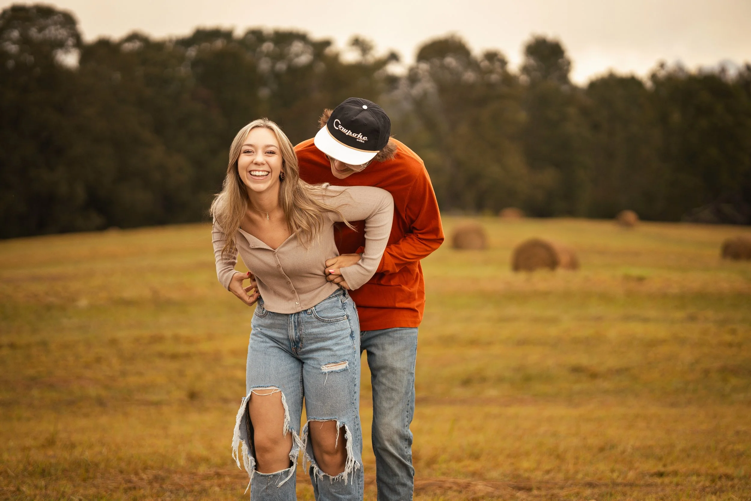 A young woman with long blonde hair and ripped jeans smiling and leaning forward, while a young man with curly hair and a black cap labeled 'Campbake' is playfully hugging her from behind in an open field with hay bales and trees in the background.