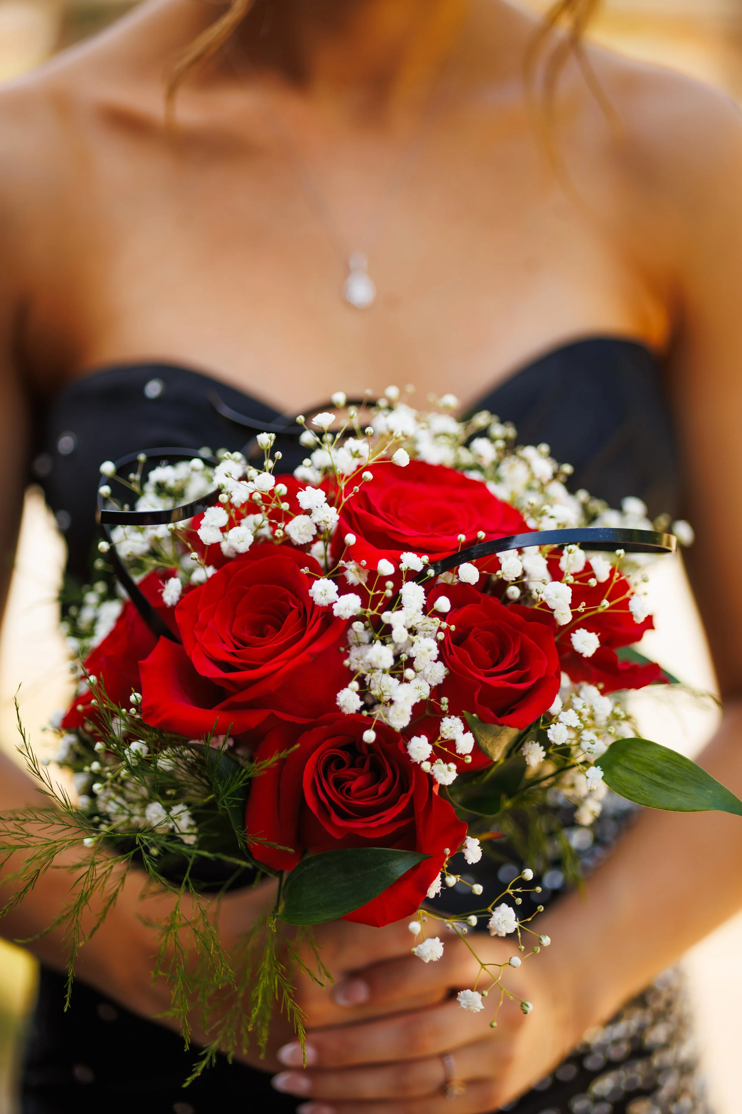 A woman in a black strapless dress holding a bouquet of red roses and white baby's breath flowers, with a pearl necklace visible on her chest.