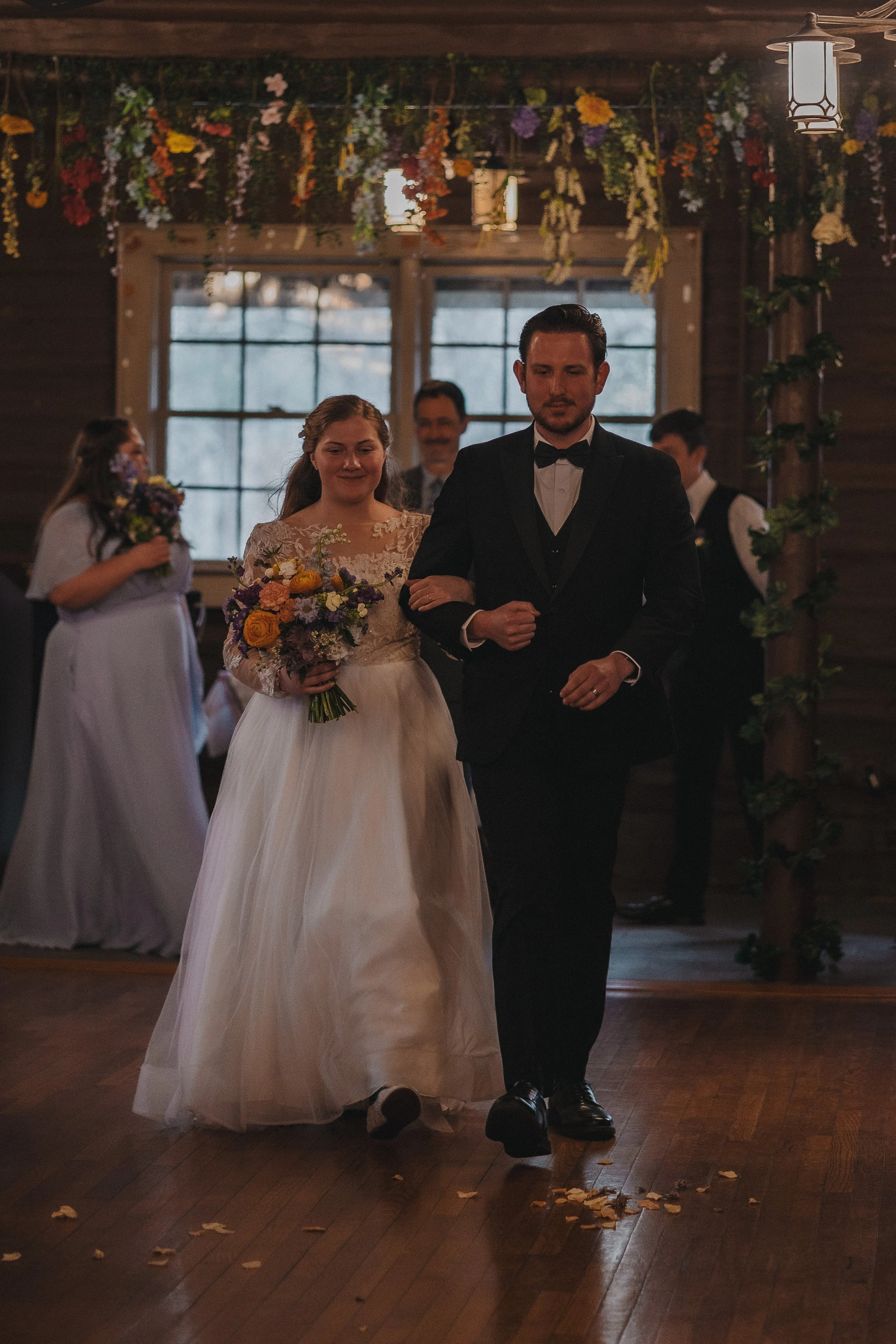 A bride and groom walking down the aisle during a wedding ceremony. The bride is holding a bouquet and wearing a lace wedding dress, while the groom is in a black tuxedo. There are bridesmaids and groomsmen in the background, with flowers and tree-li