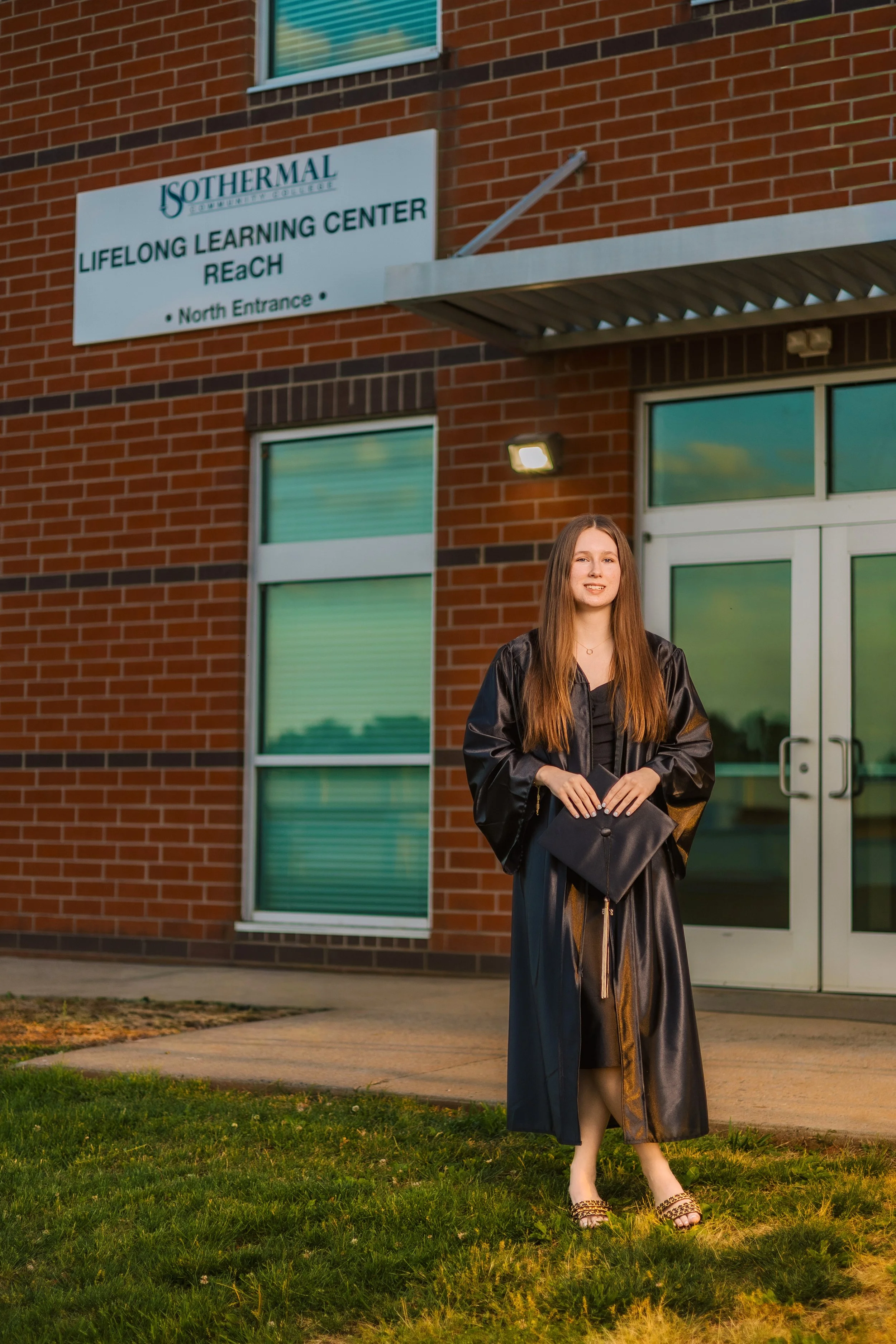 Young woman in a black graduation gown and cap standing outside in the grass, in front of a brick building with green-tinted windows and a white sign that reads ISOTHERML Summer Institute, Lifelong Learning Center, REACH, North Entrance.