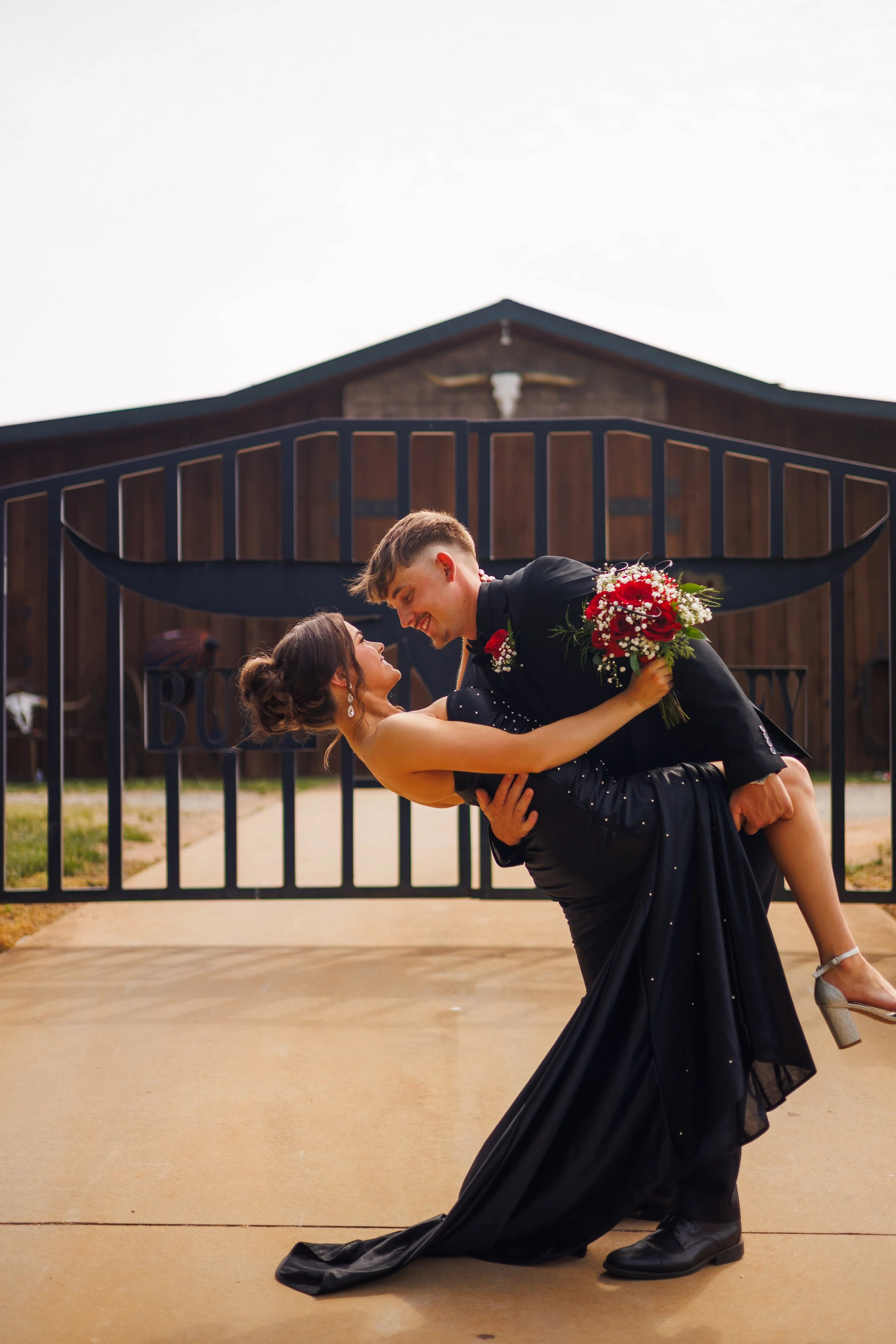 A couple celebrating a wedding outdoors, with the groom lifting the bride who is holding a bouquet of red and white flowers, in front of a barn-style building.