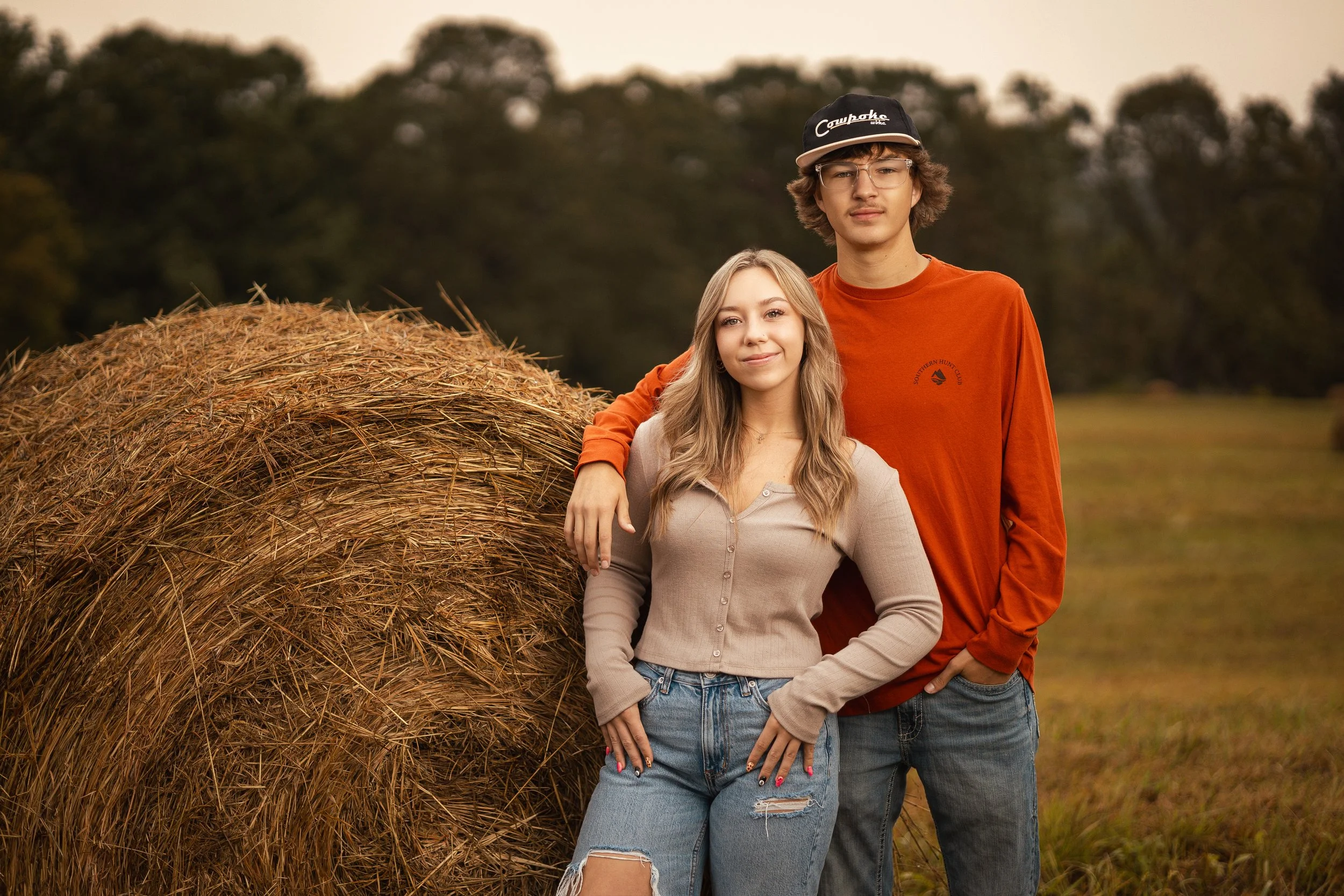A young woman and a young man standing side by side outdoors next to a large hay bale with a wooded background. The woman has long blonde hair, is smiling slightly, and is wearing a beige button-up sweater and ripped jeans. The man has curly brown ha