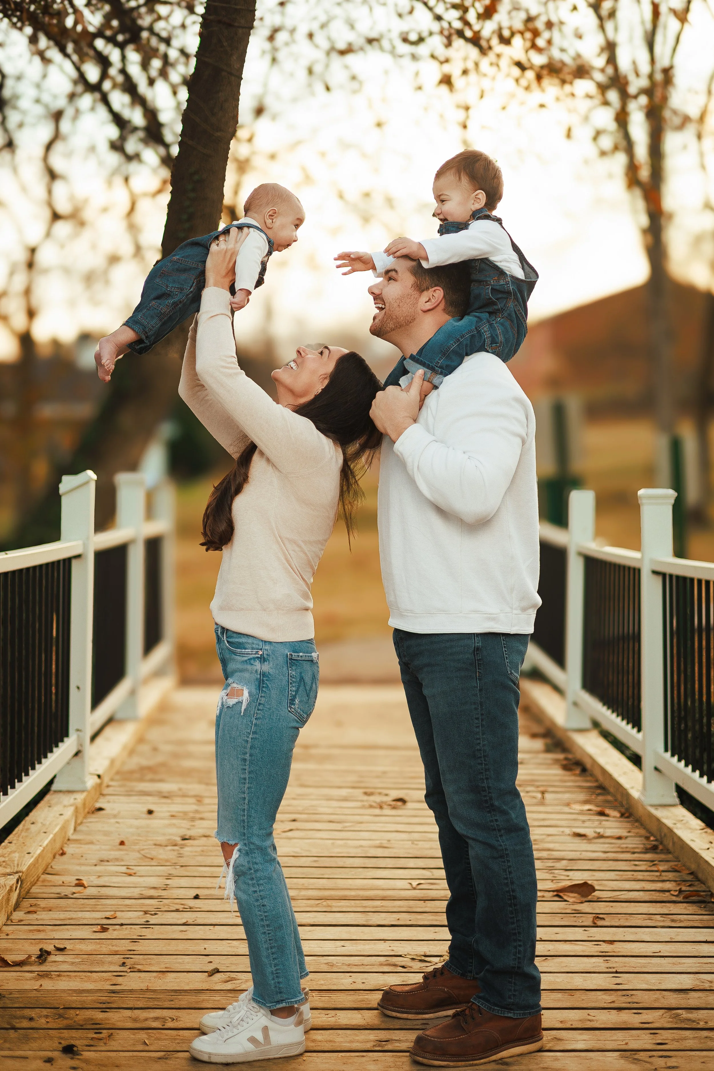 A family of four outdoors on a wooden bridge. The father has a boy on his shoulders, reaching out to another boy in front. The mother lifts the boy in front, and they all smile at each other. The background has trees with sunlight filtering through.