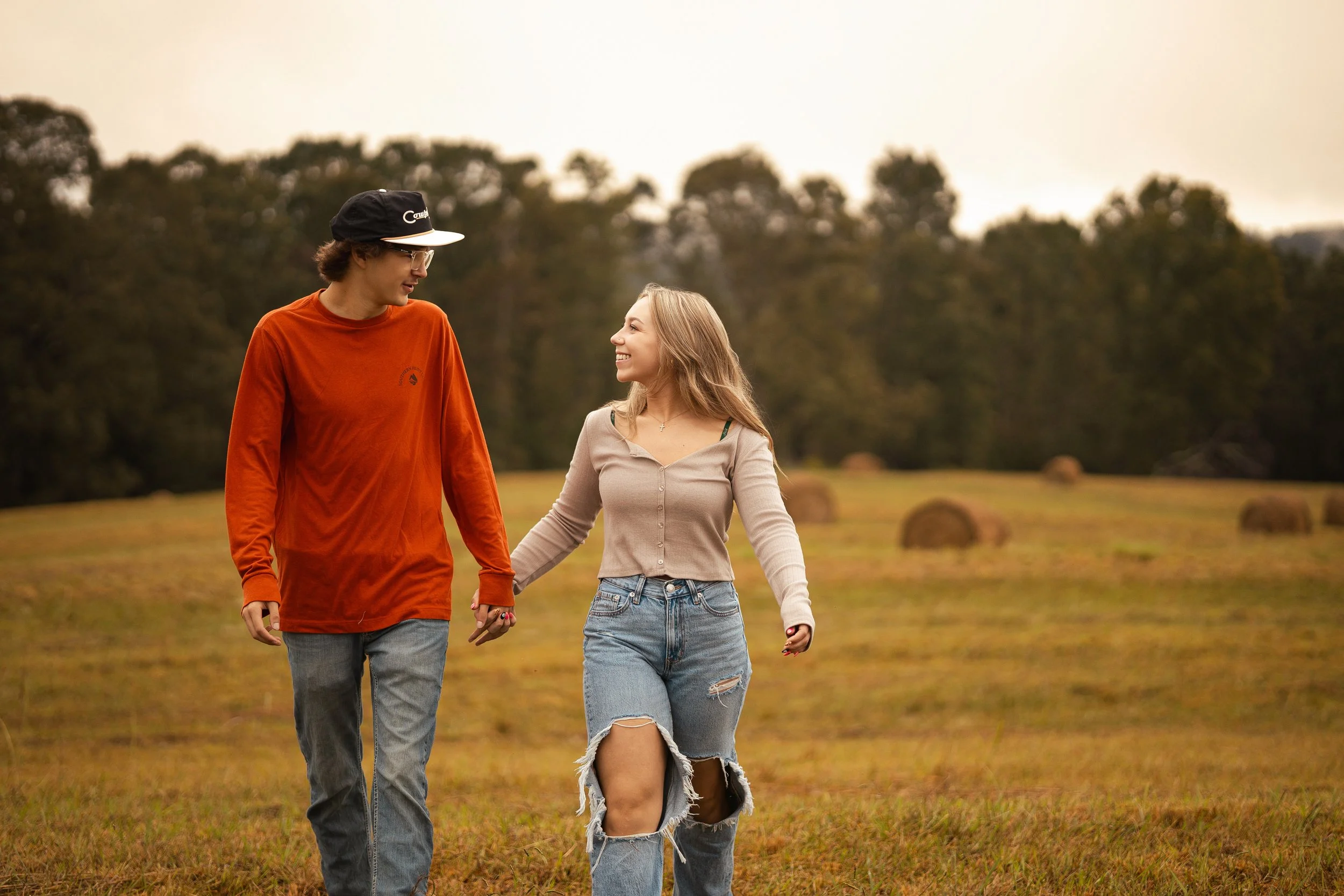 A young couple holding hands and walking in a field with hay bales, with trees and an overcast sky in the background.