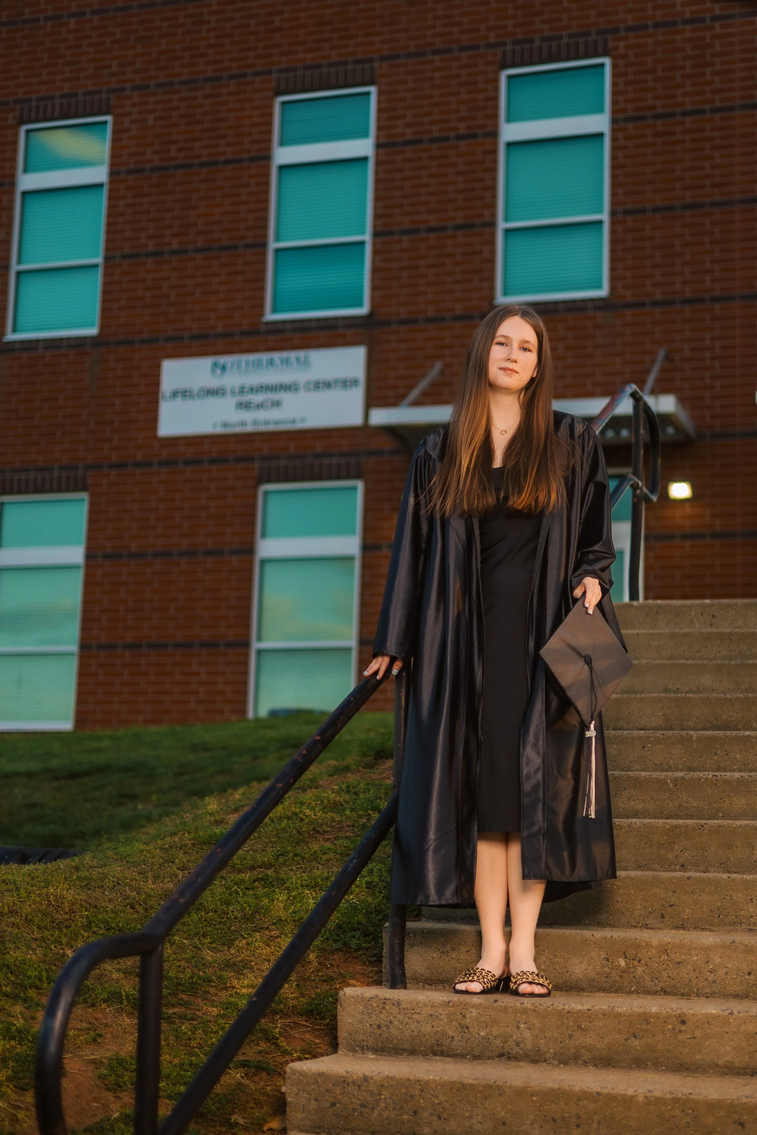 A young woman in a black graduation gown holding a black cap, standing on concrete stairs outside a brick building with green windows.