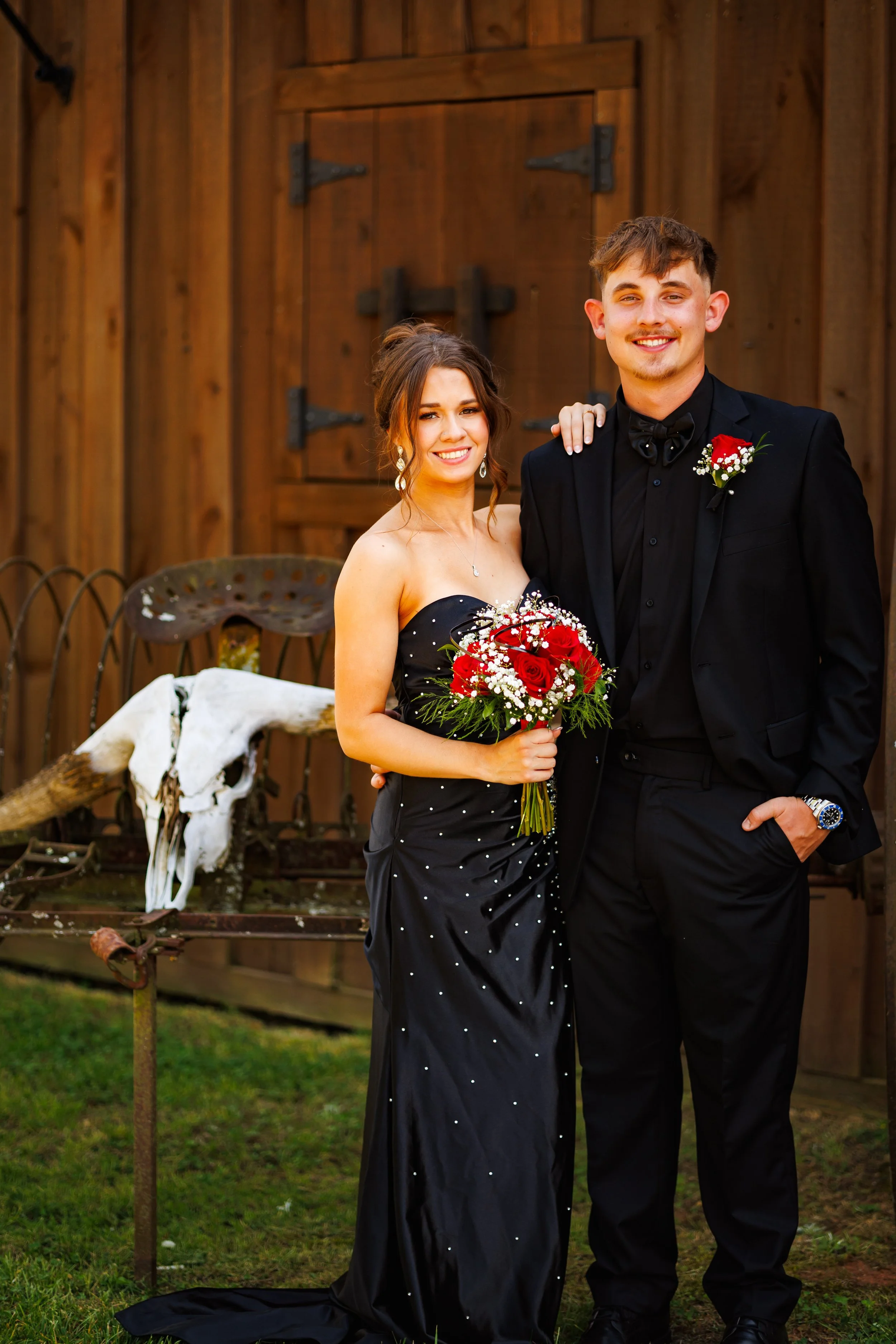 A young man and woman dressed in formal attire, standing outdoors in front of a wooden barn, with the woman holding a bouquet of red roses and wearing earrings, while the man has a boutonnière on his jacket.