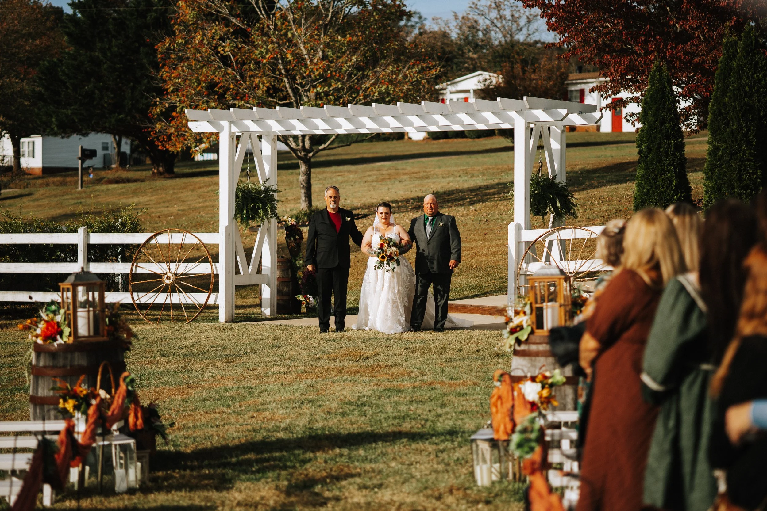 A bride walking with two men under a white garden arch at an outdoor wedding ceremony, with autumn trees and chairs with guests visible in the background.