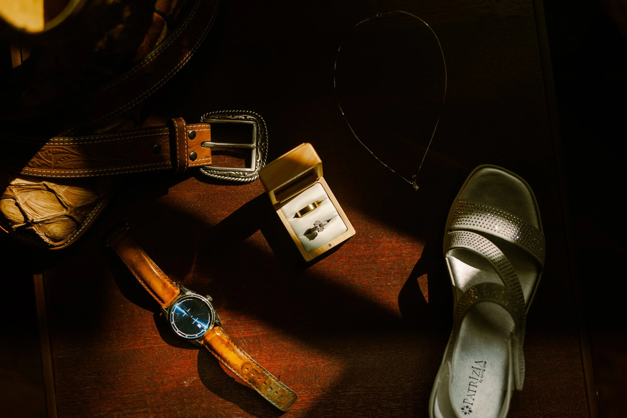 A wristwatch with a brown leather strap, a pair of white sneakers with rhinestones, a jewelry box with an engagement ring, a silver necklace, a brown leather belt, a black handbag, and part of a straw hat on a wooden surface.