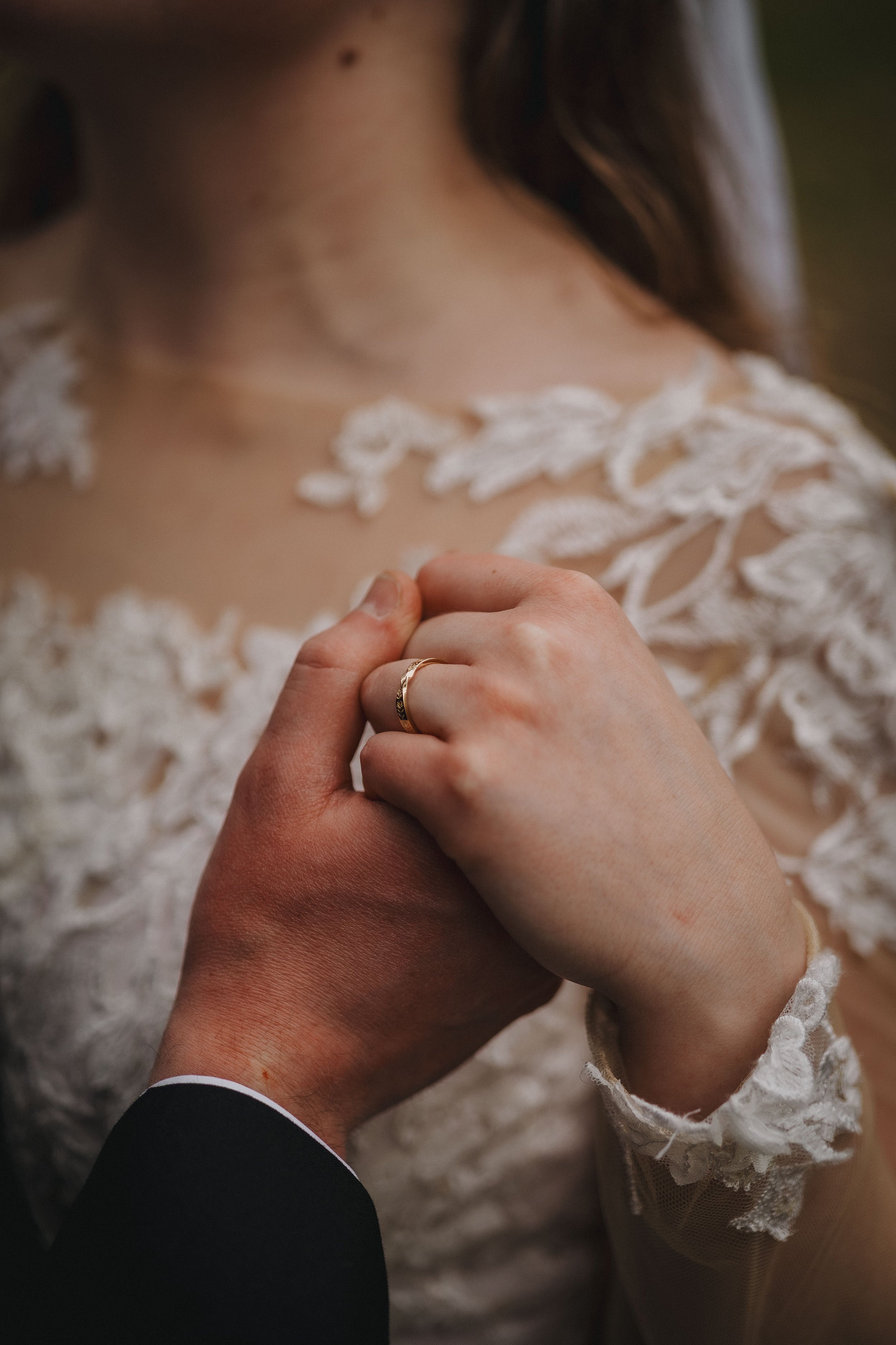 Close-up of a man and woman holding hands during a wedding. The woman's hand has a ring, and she is wearing a wedding dress with lace details. The man's hand is partially visible, and he is wearing a dark suit.