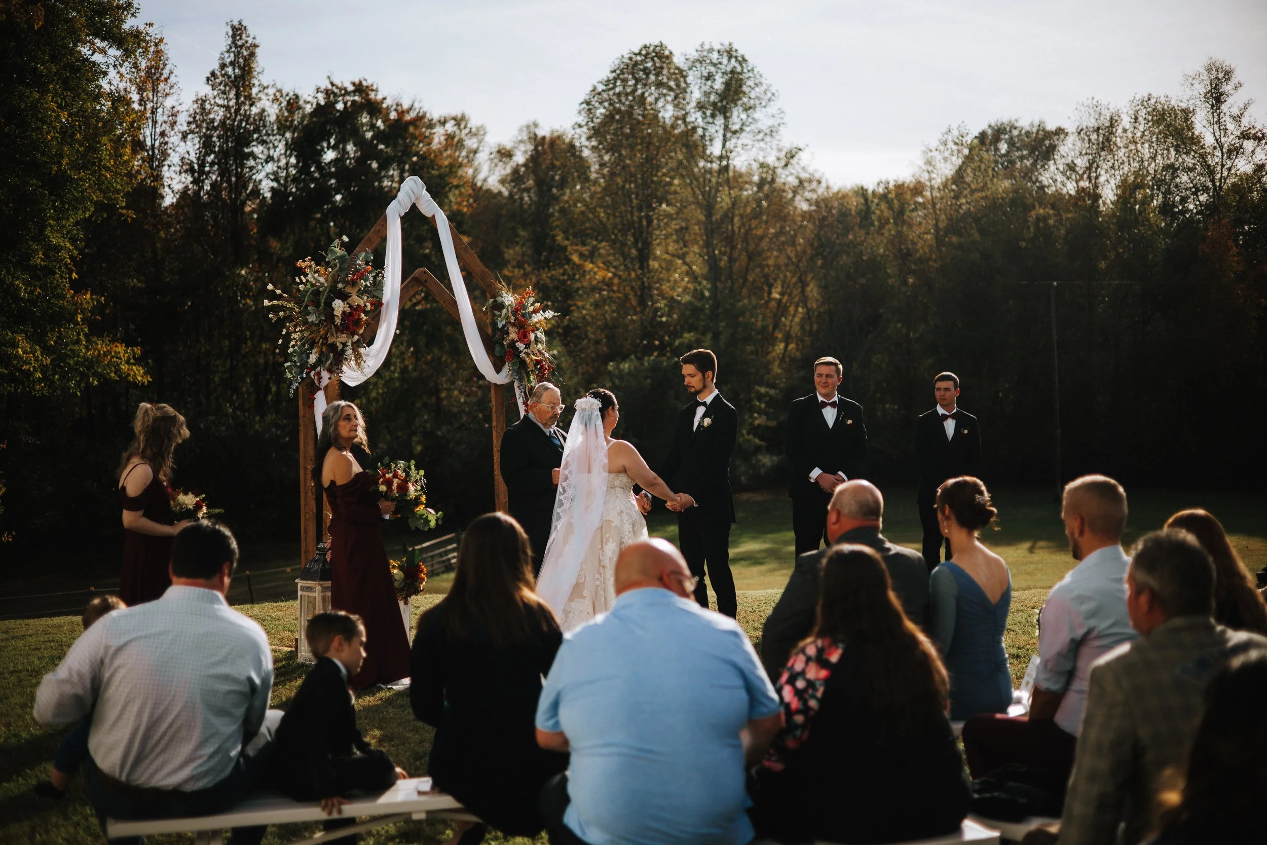 A outdoor wedding ceremony with a bride and groom holding hands, officiant, bridesmaids, groomsmen, and guests seated on grass, under a decorated arch, during sunset with trees in background.