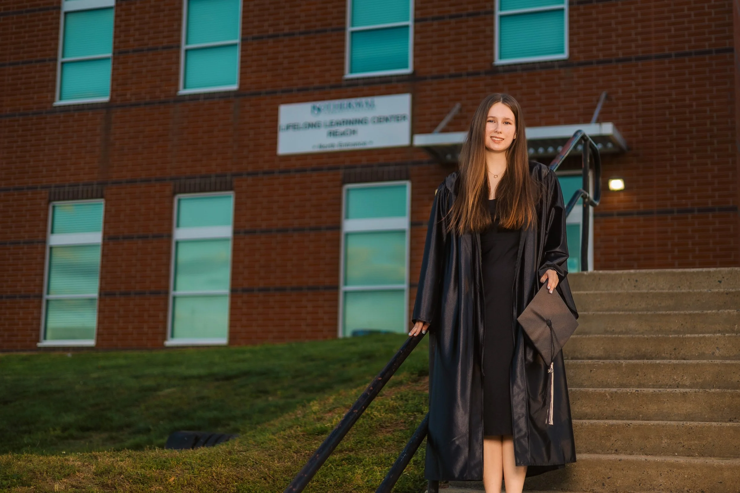 A young woman in a black graduation gown and cap holding a diploma stands on steps outside a brick building with a sign reading "Lifelong Learning Center."