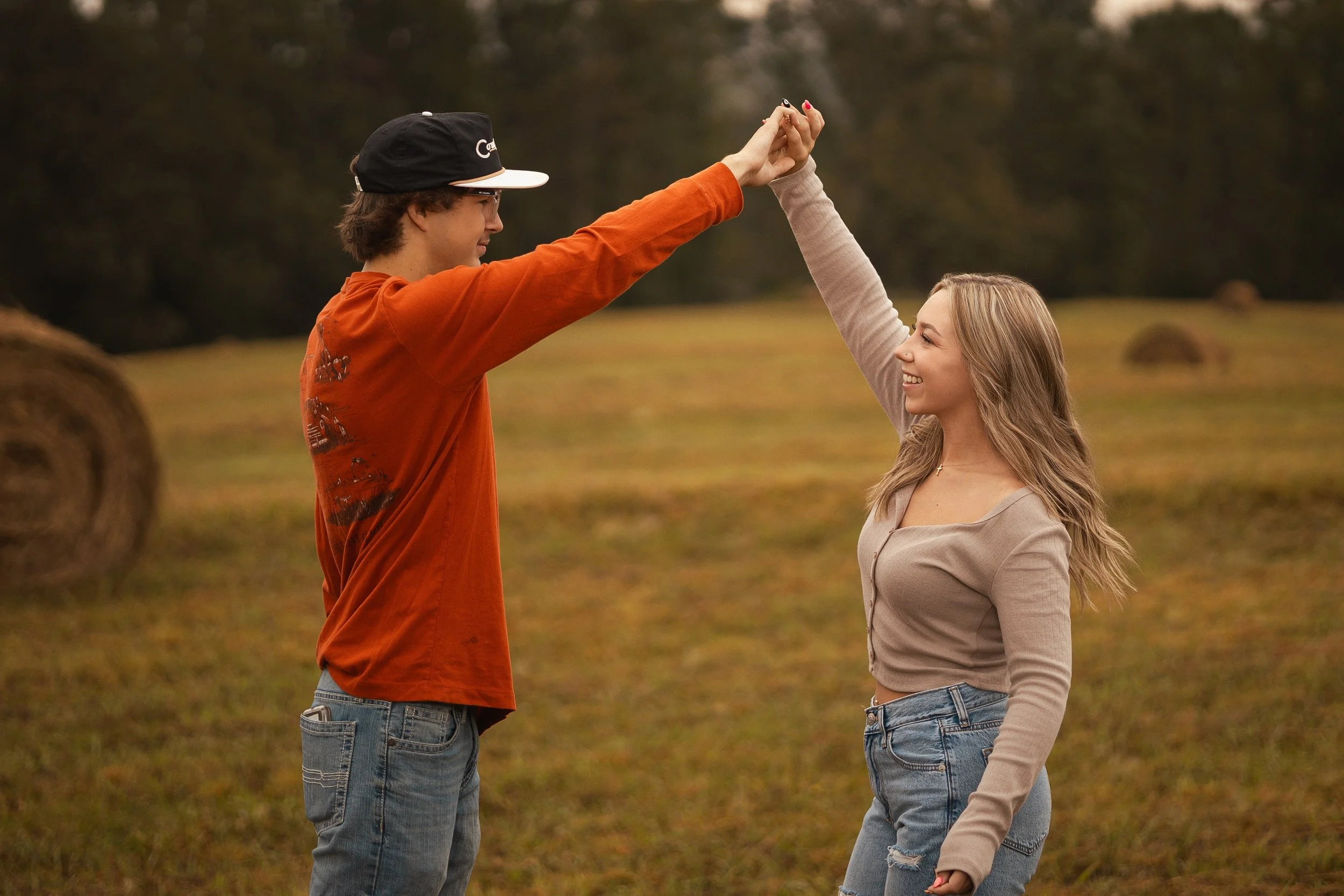 A young man and woman stand outdoors in a field with hay bales, smiling and holding hands up together in a celebratory gesture as they look at each other.