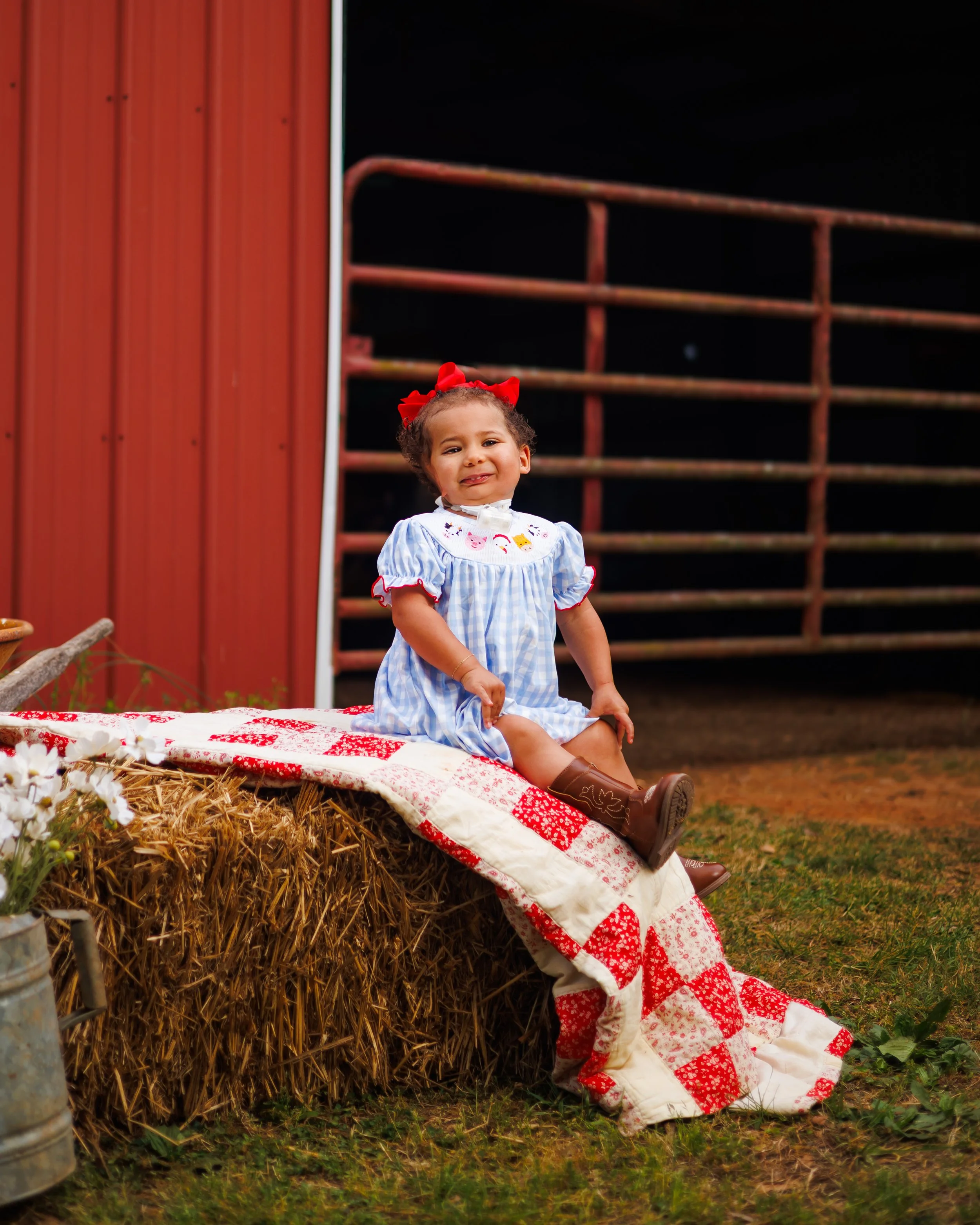 Young girl with curly hair, red bow, and dress with a pig pattern, smiling while sitting on a quilt on a hay bale in a farm setting at night.