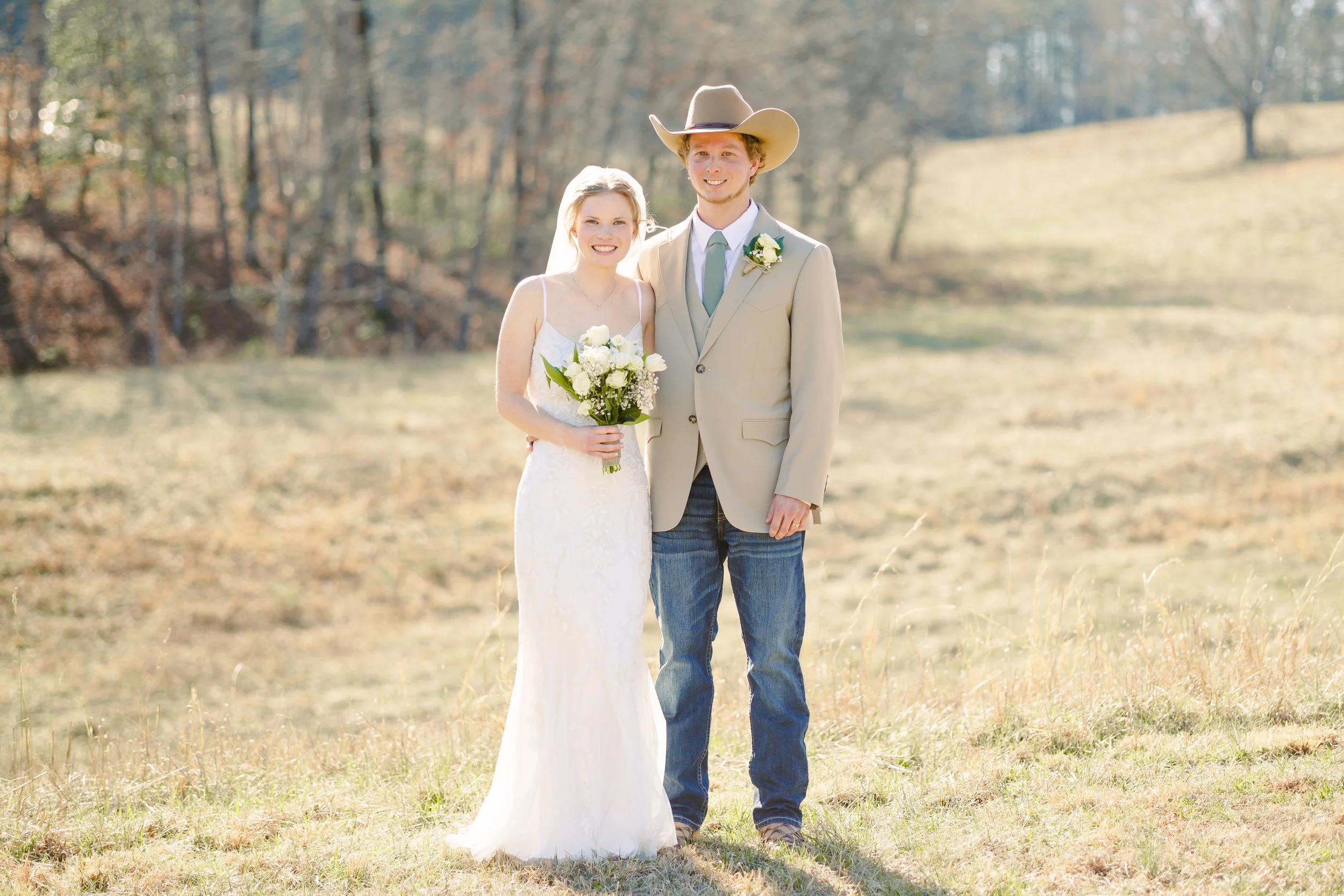 A newlywed couple standing outside in a grassy field with trees in the background, smiling at the camera. The bride is holding a bouquet of white flowers, wearing a white wedding dress. The groom is dressed in a beige suit with a light blue tie and a