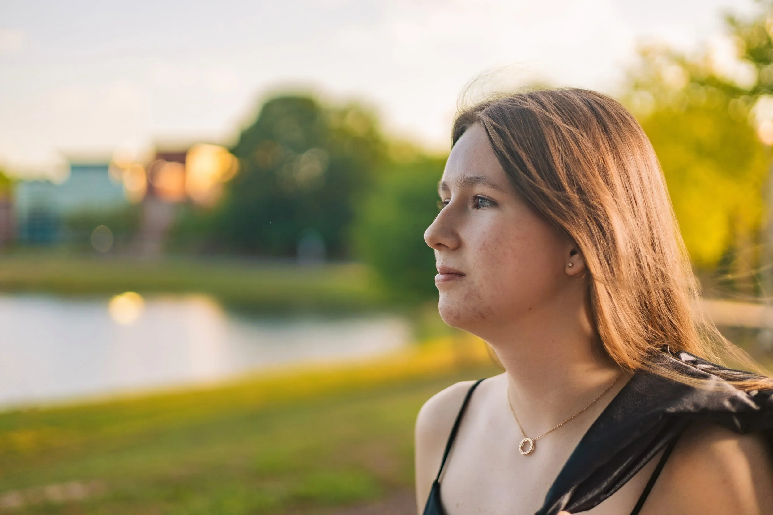 A young woman with red hair and a small earring looks to the right outdoors during sunset, with blurred trees and a lake in the background.