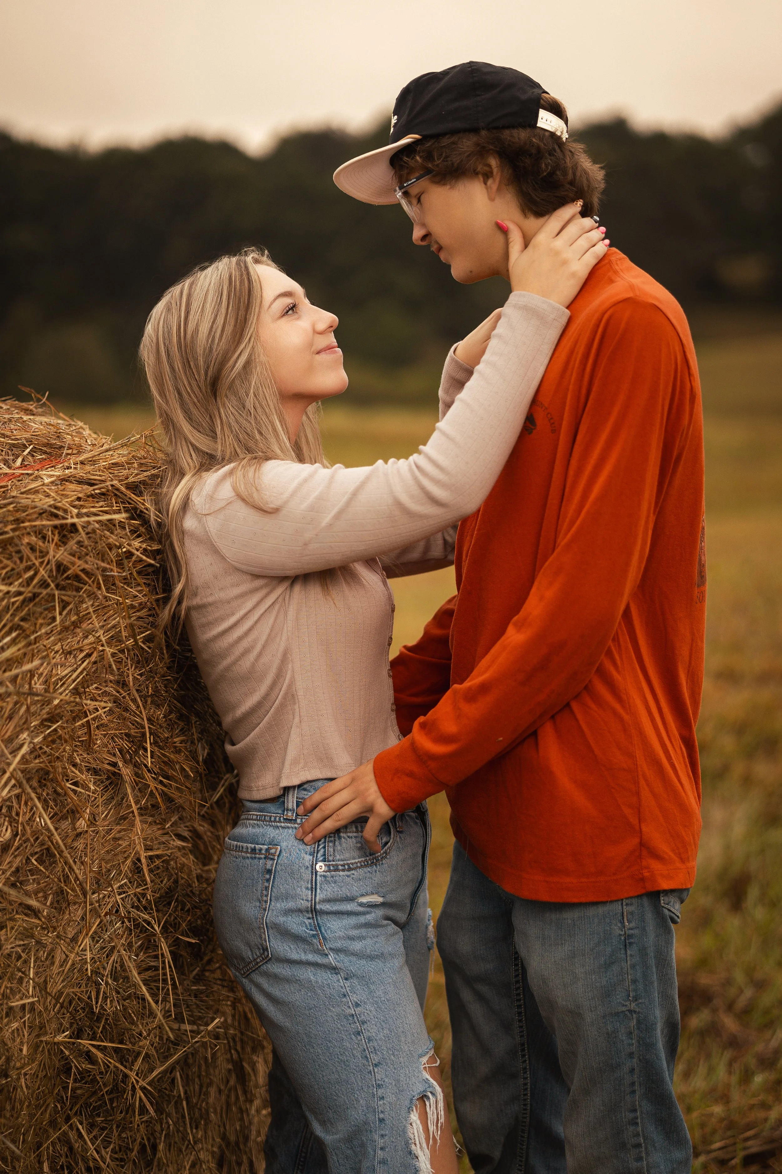 A young woman with blonde hair and a beige sweater holding a young man with glasses, a black cap, and an orange shirt close to her in an outdoor field with a haystack and trees in the background.