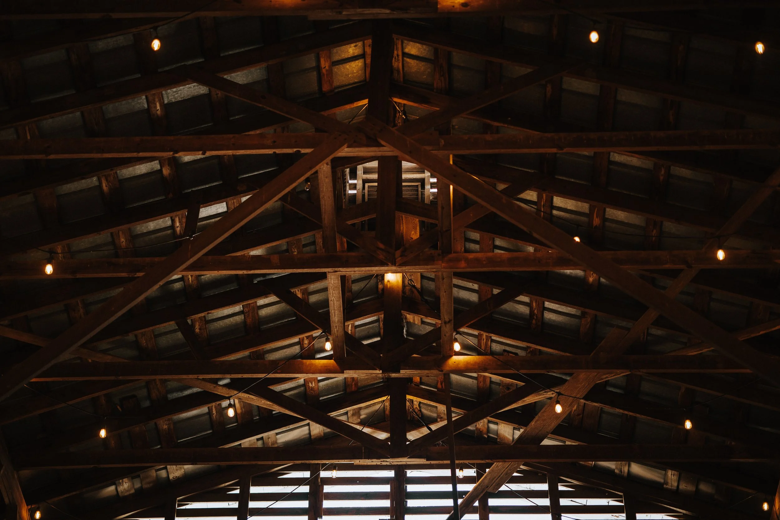 Looking up at the wooden rafters and beams of a barn or attic ceiling, illuminated by small hanging light bulbs.