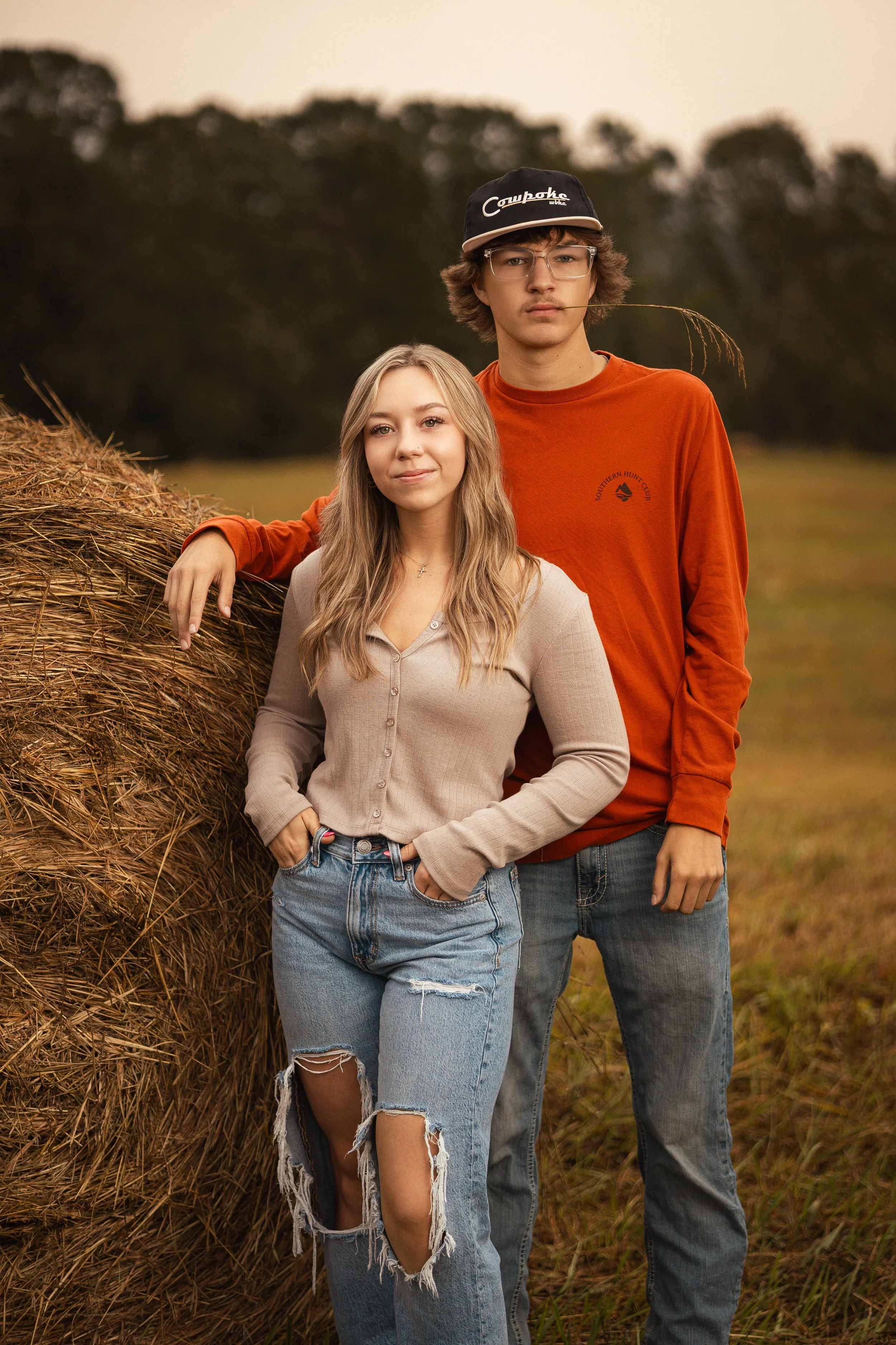 A young woman leaning against a hay bale and a young man standing beside her in an outdoor rural setting with trees in the background at sunset.