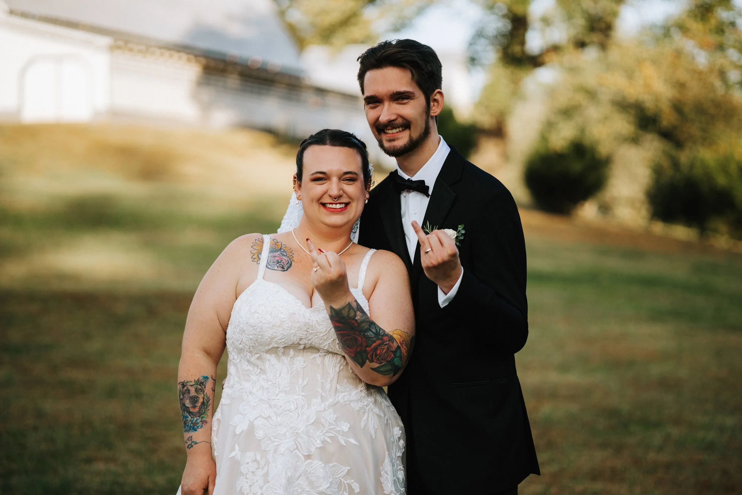A smiling couple in wedding attire standing outdoors on a sunny day, holding up their middle fingers.