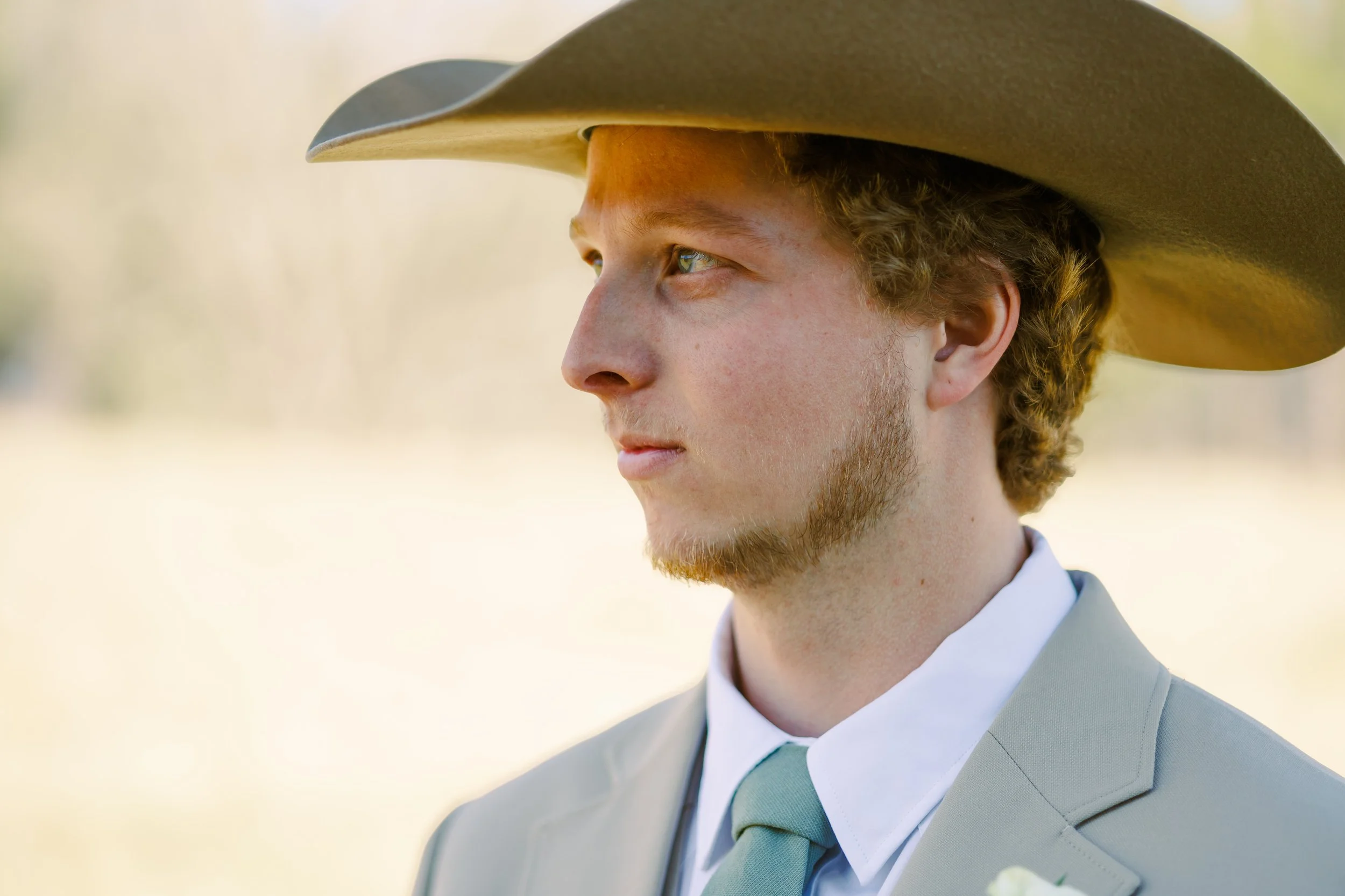 A young man with light skin and curly reddish-brown hair wearing a gray suit, white shirt, tie, and a gray hat, looking to the left outdoors with a softly blurred natural background.