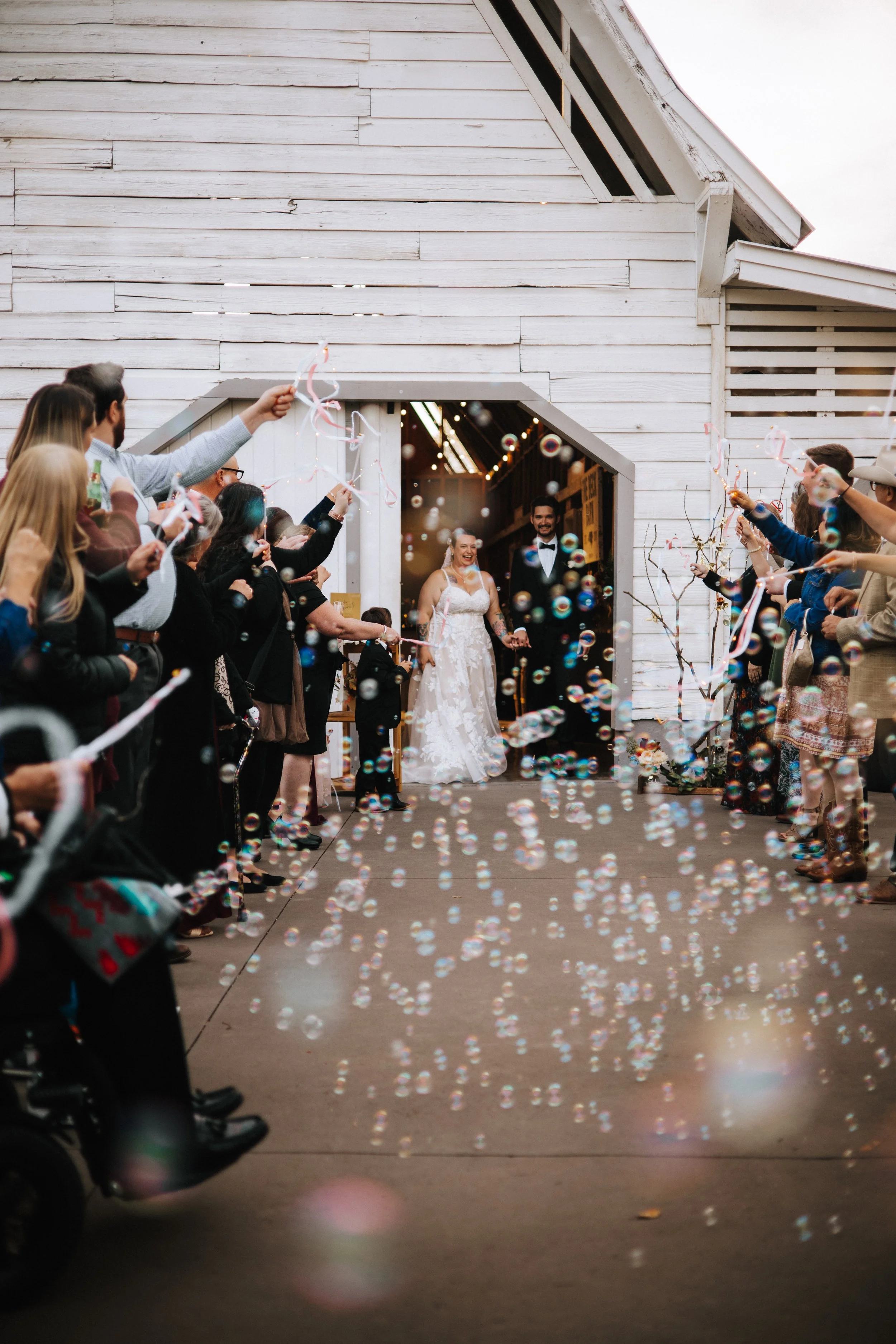 A bride and groom smile as they walk out of a rustic barn, surrounded by guests and children celebrating with bubbles and party streamers.