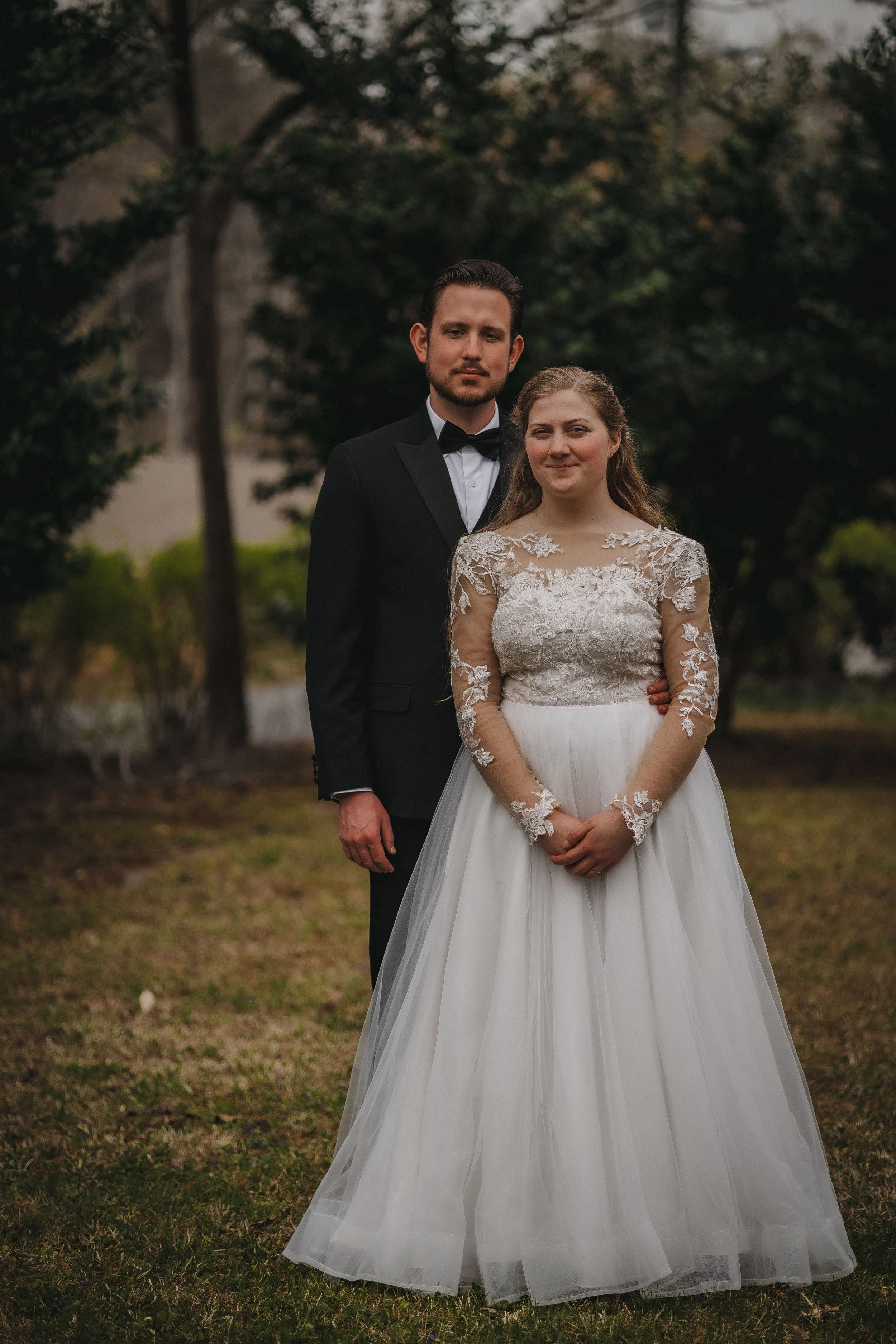 A couple in wedding attire standing outdoors, with the groom in a black tuxedo and the bride in a white wedding dress with lace details. They are posing in a park with trees in the background.