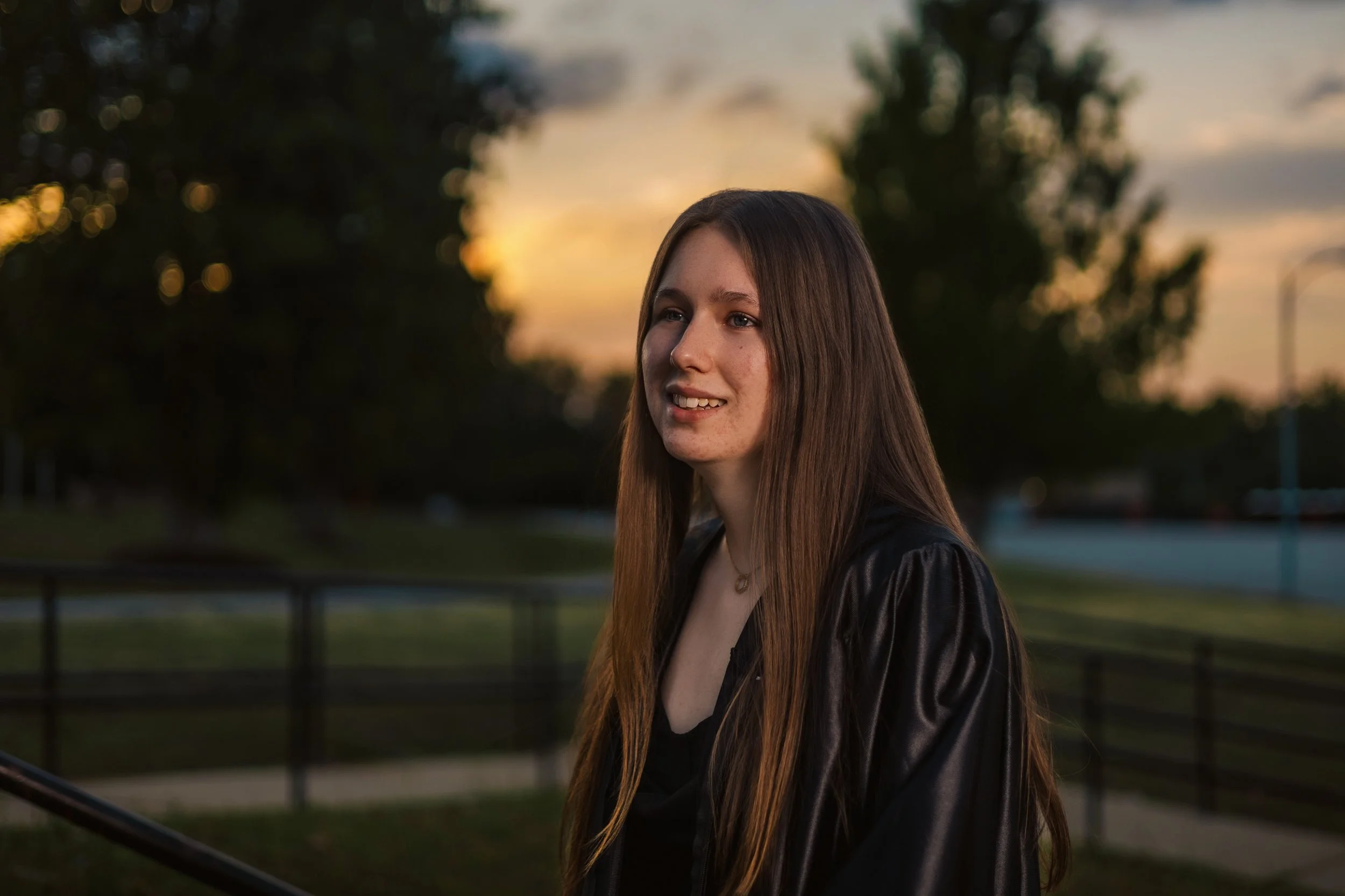 A young woman with long brown hair standing outdoors during sunset, smiling gently, wearing a black leather jacket.