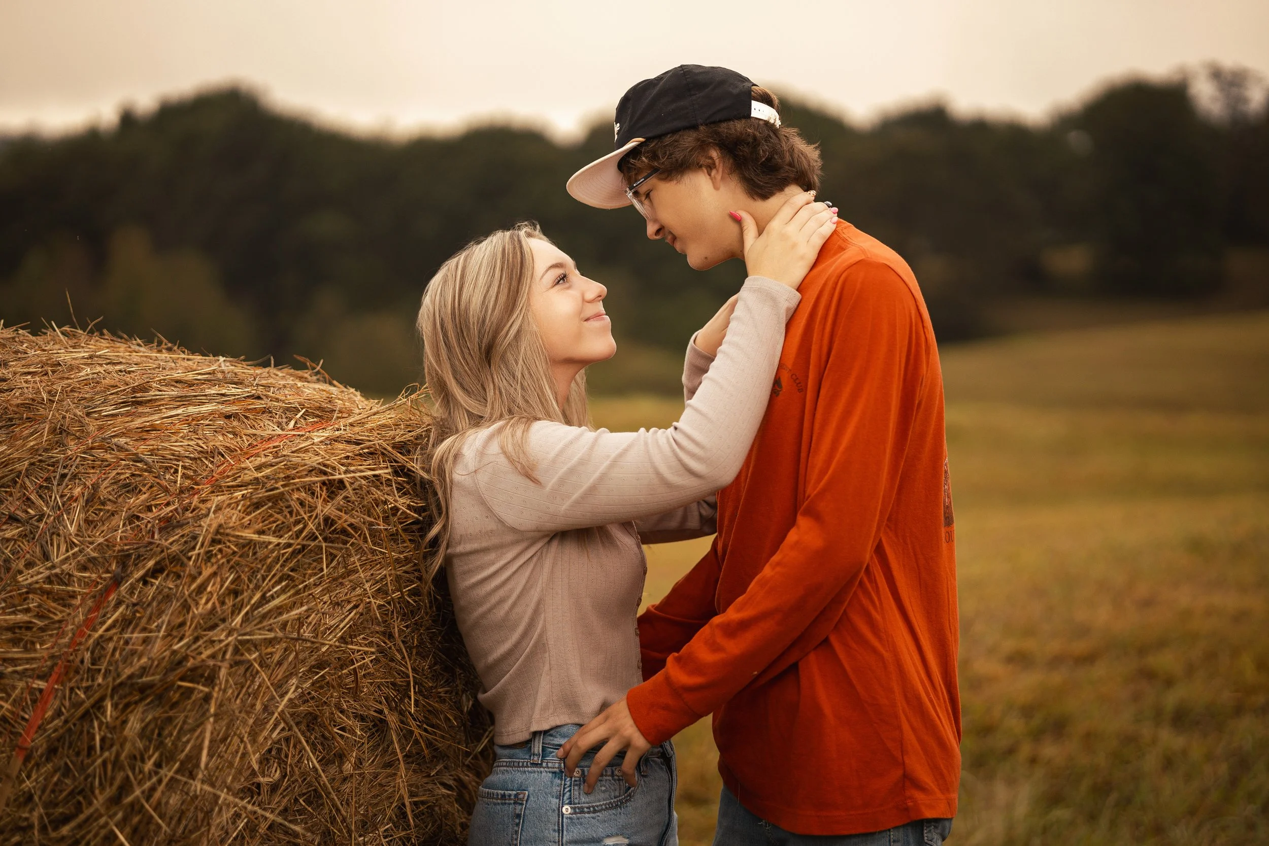 A young woman and young man share an intimate moment outdoors near a hay bale, with the woman holding the man's neck and looking up at him affectionately, both smiling.