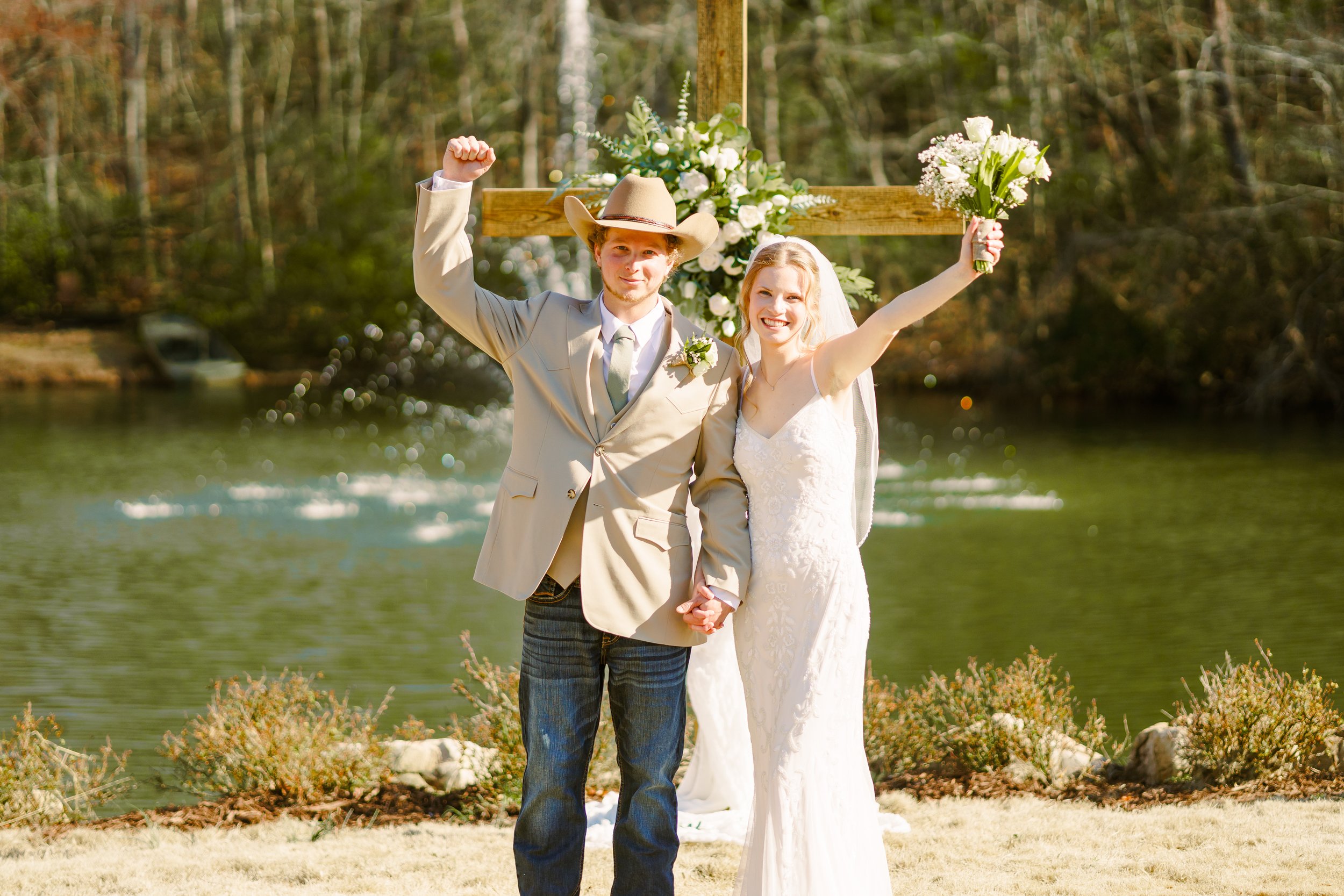 A bride and groom standing outdoors by a lake, holding hands, with a wooden cross decorated with white flowers behind them, during a wedding ceremony.