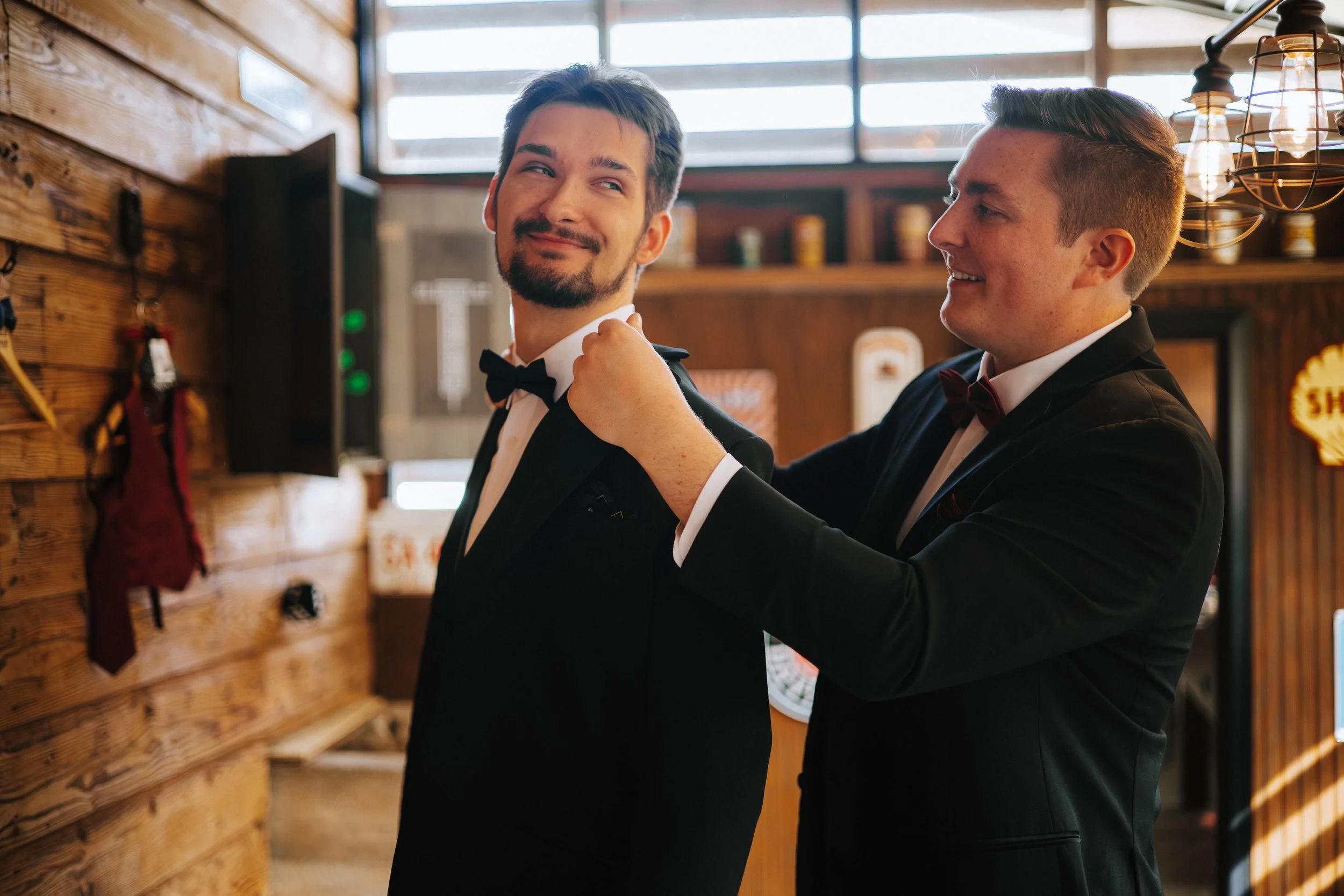 Two men in tuxedos smiling inside a rustic wooden venue, with one adjusting the other's bow tie.