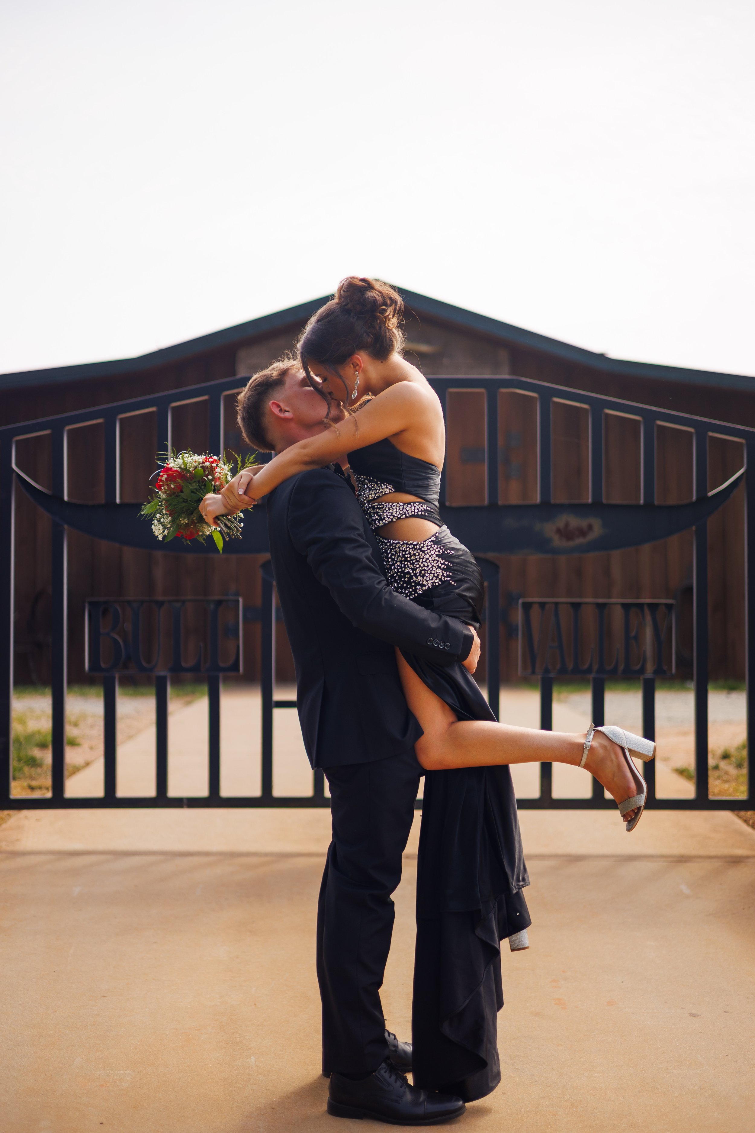 A man in a tuxedo lifting a woman in a black dress in front of a gate with the words "Bulle" and "Valley", with a barn behind them. The woman holds a bouquet of red and white flowers and is wearing silver high heels.