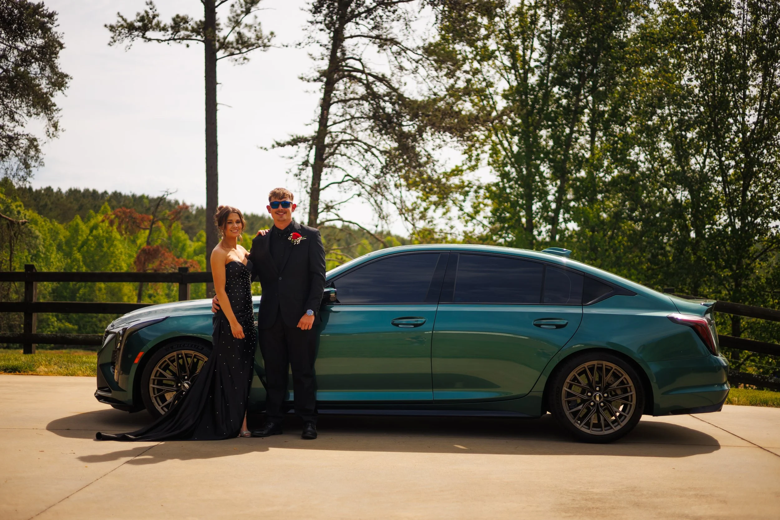 A couple in formal attire standing beside a dark green sports car outdoors, with trees and a fence in the background.