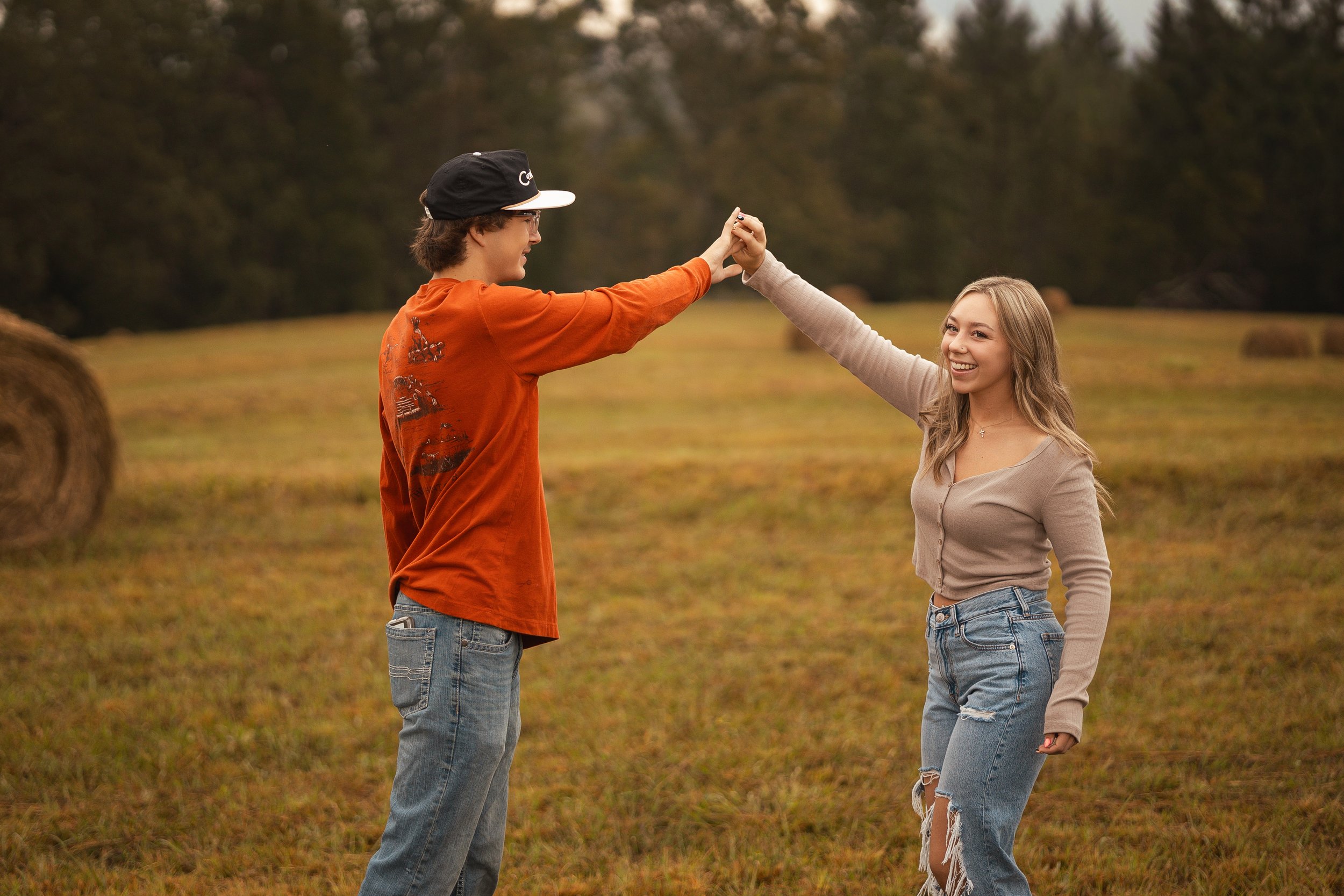 A young man and woman are outdoors in a field with hay bales, holding hands and smiling at each other.