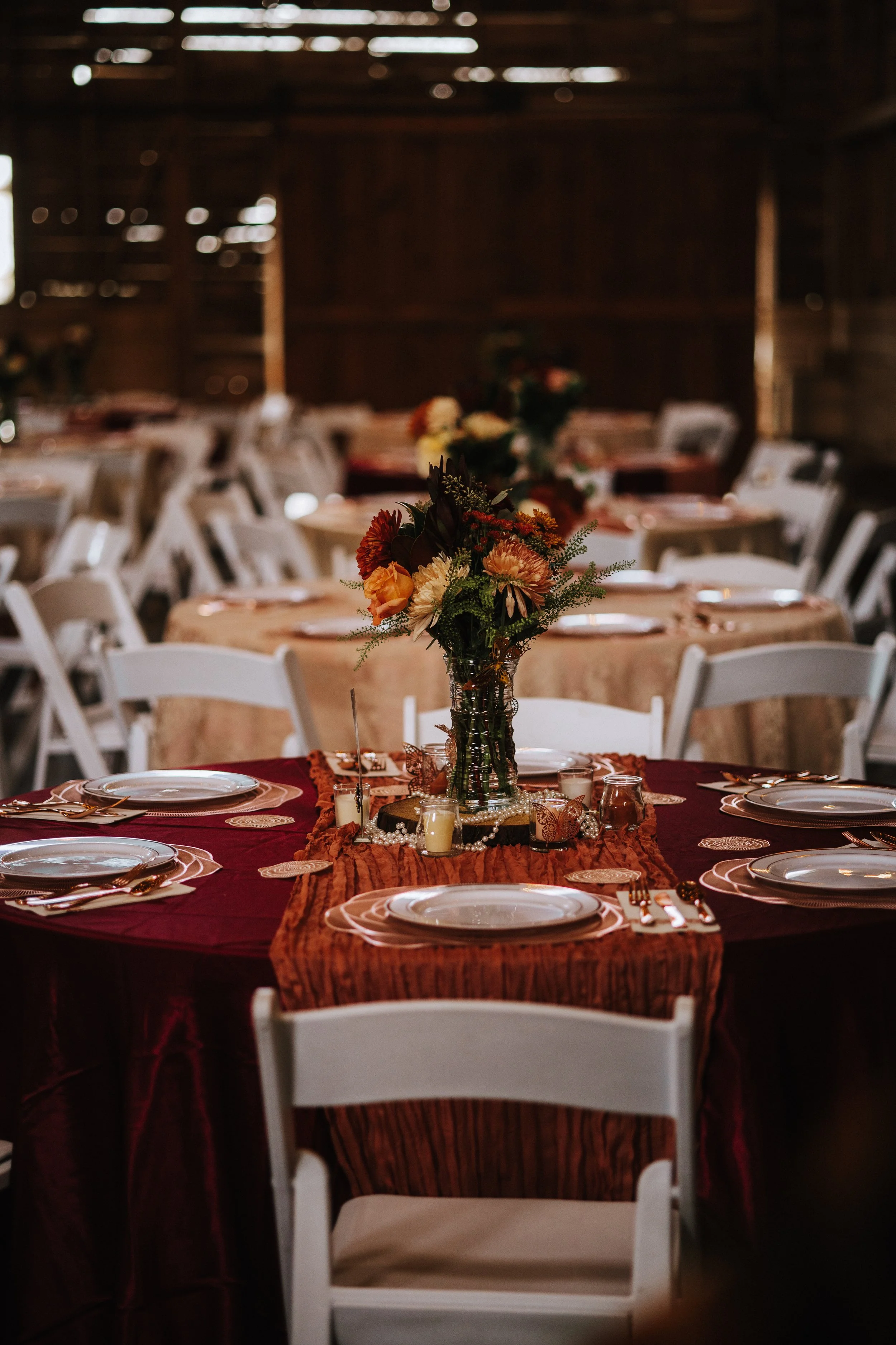 A banquet table is decorated with a bouquet of orange, peach, and cream-colored flowers in a glass vase. The table is covered with a burgundy tablecloth and ruffled orange runner. Place settings include white plates, silverware, and napkins with deco