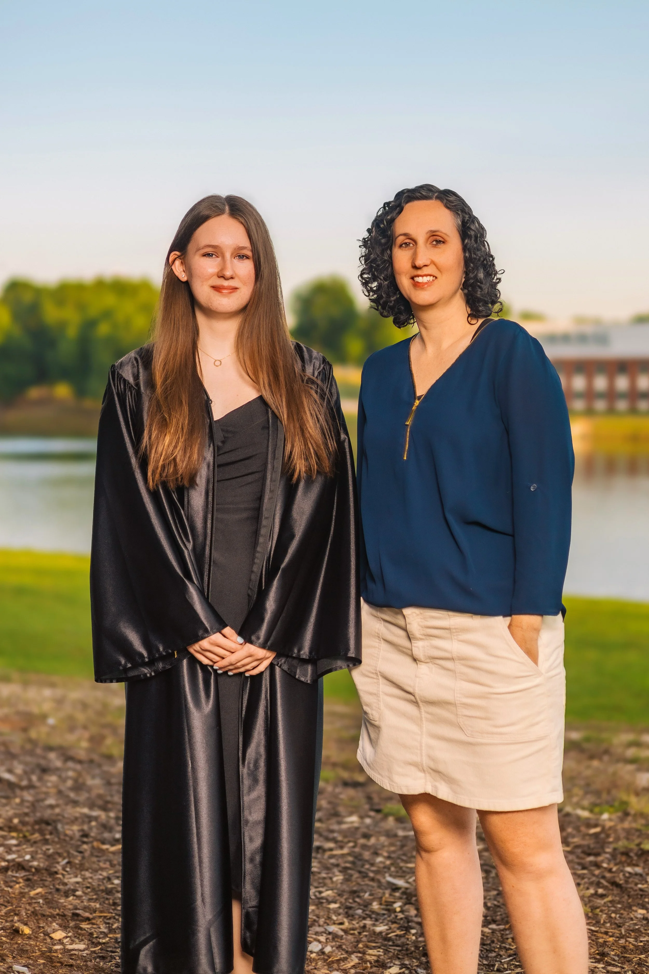 Two women standing outdoors near a body of water with trees and a building in the background, one in a graduation gown and the other in casual beige shorts and a blue blouse.