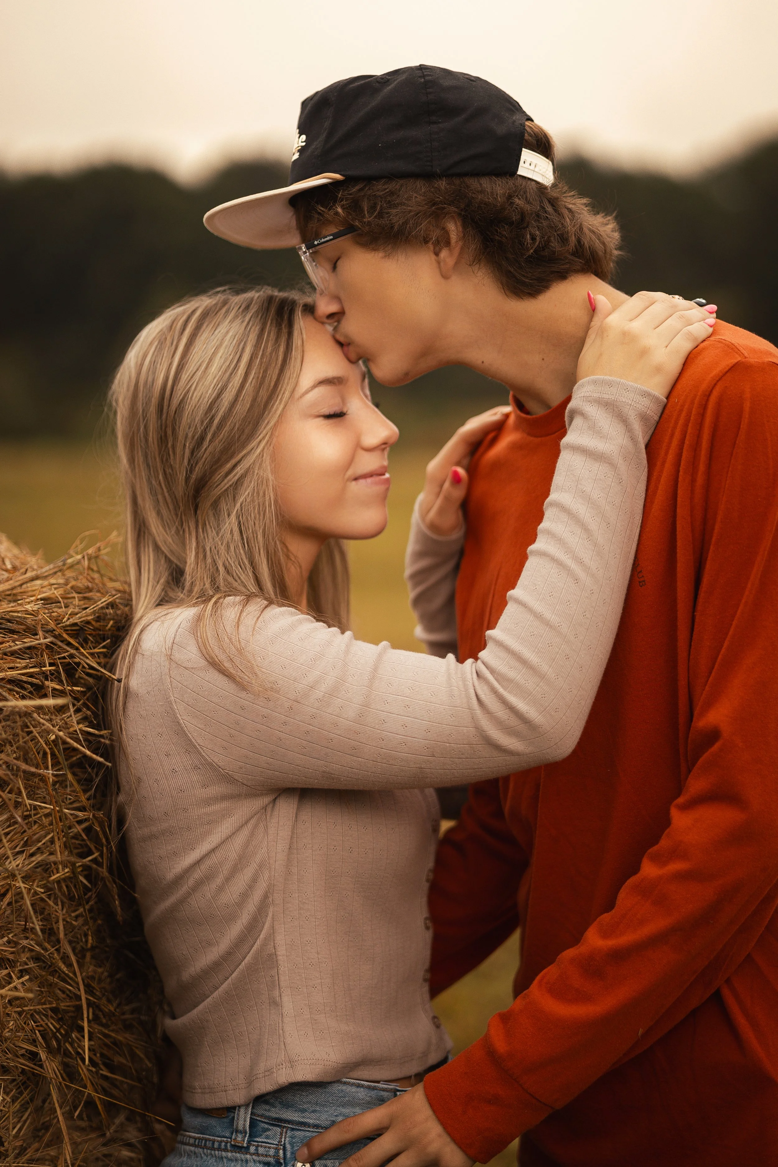 A young couple embraces outdoors, with the man kissing the woman's forehead and holding her face gently. The woman has blonde hair and her eyes closed, and she smiles softly. The background is blurred with greenery.